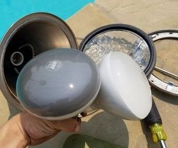 A pool repair person is holding a pool light bulb in their hand in front of a pool.