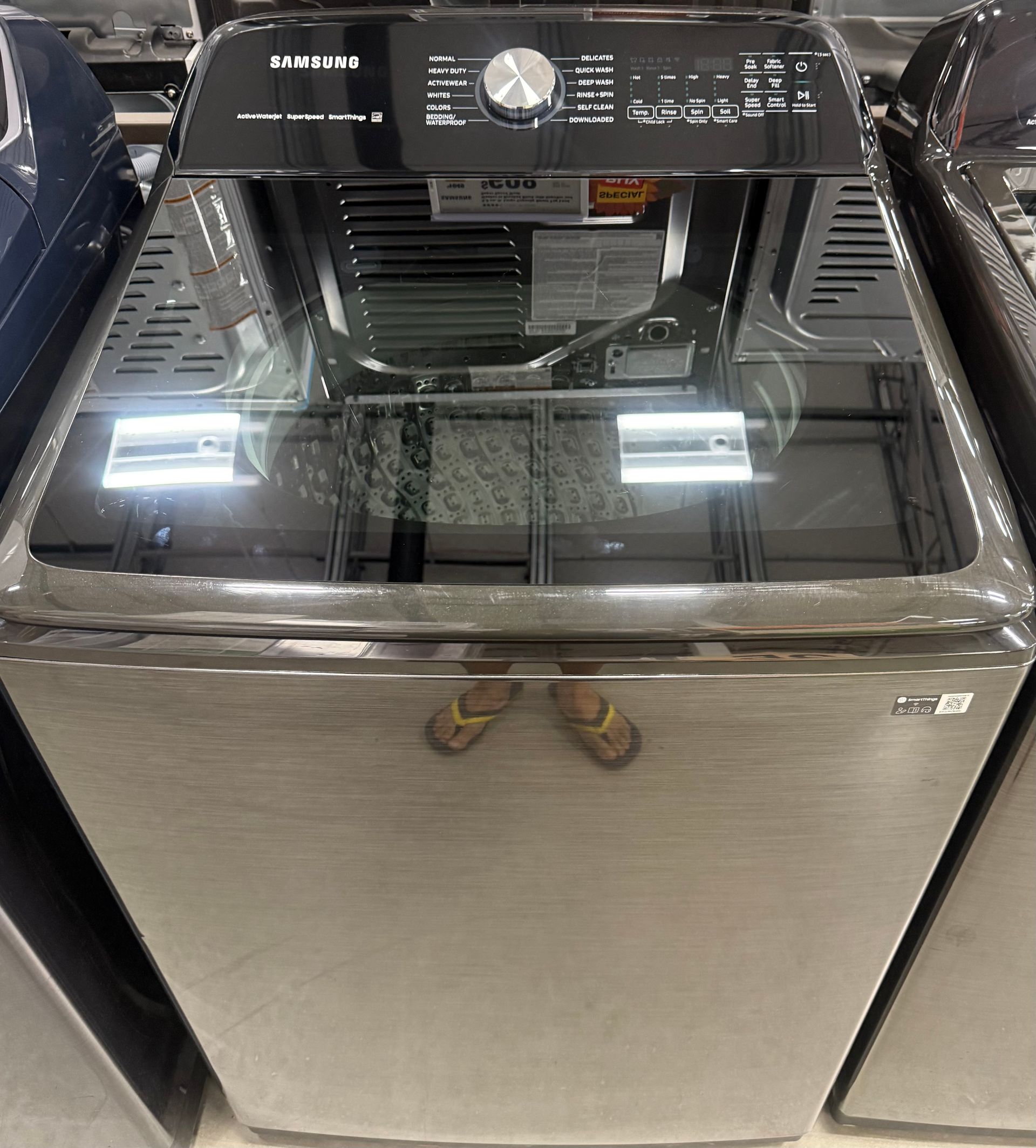 Samsung top-load washing machine, stainless steel finish, with flip-flops resting on the glass lid.