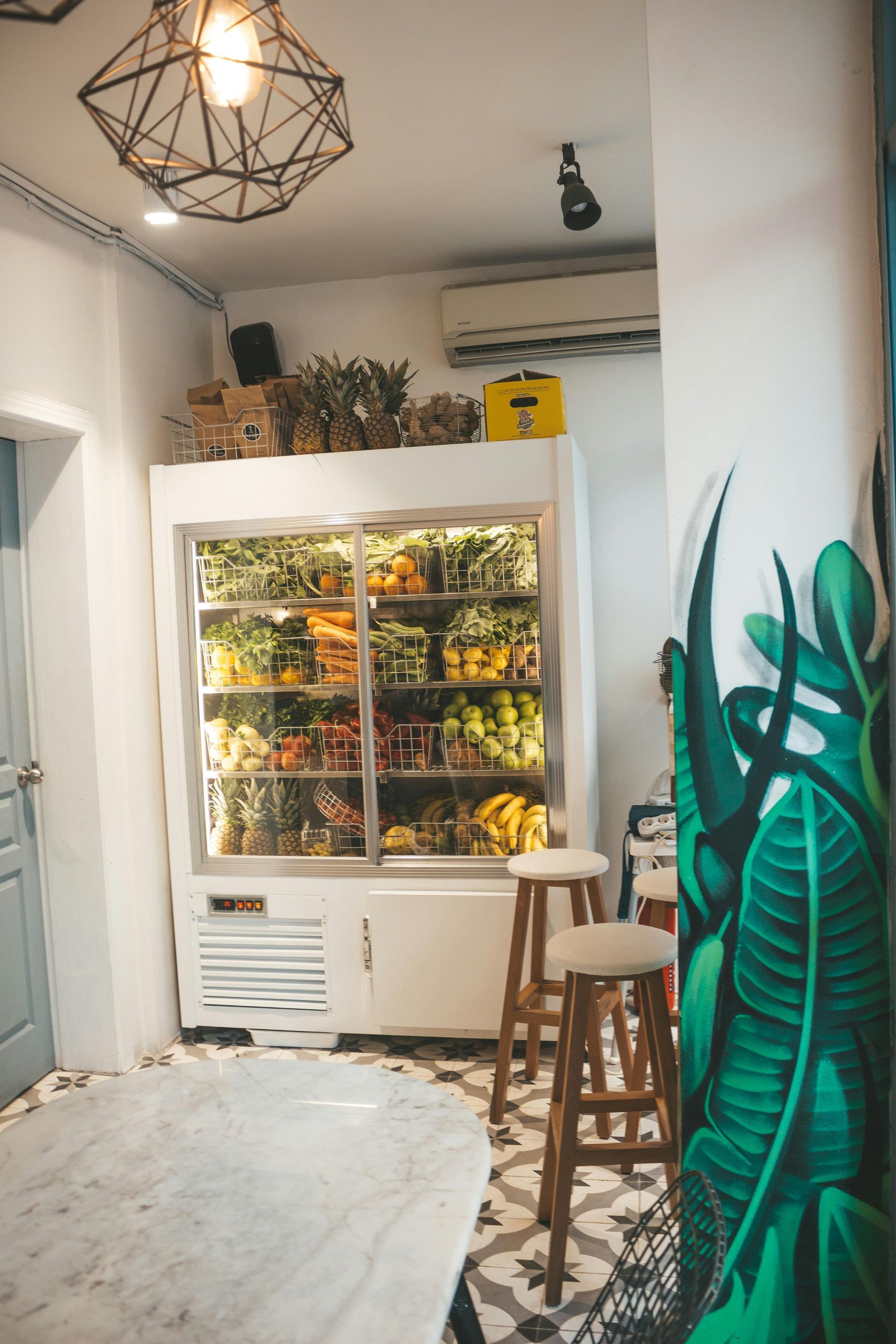 A refrigerator filled with colorful fruits and vegetables in a brightly lit cafe, stools nearby.