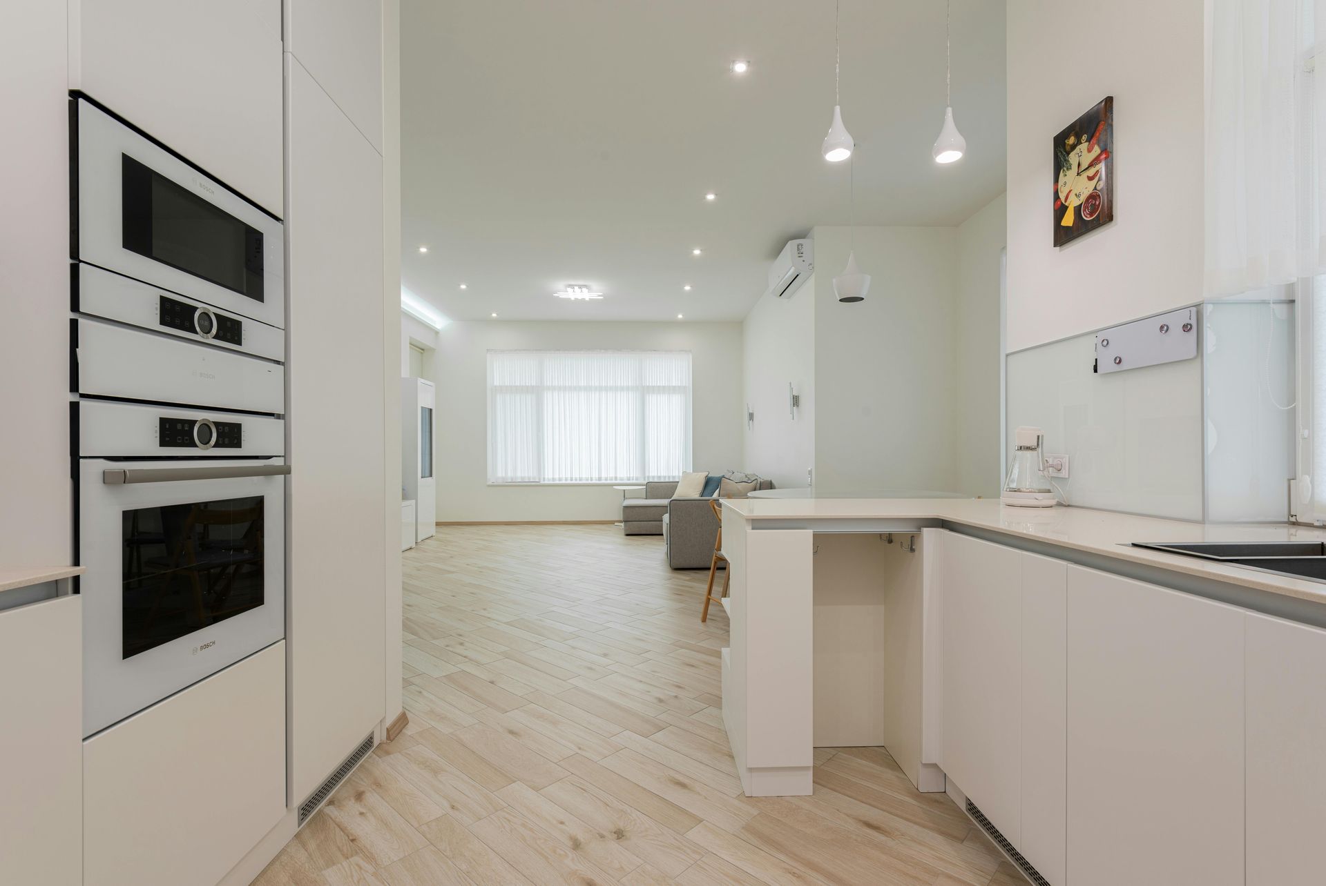 White kitchen with oven and microwave. Open to a bright living room with windows and sofa.