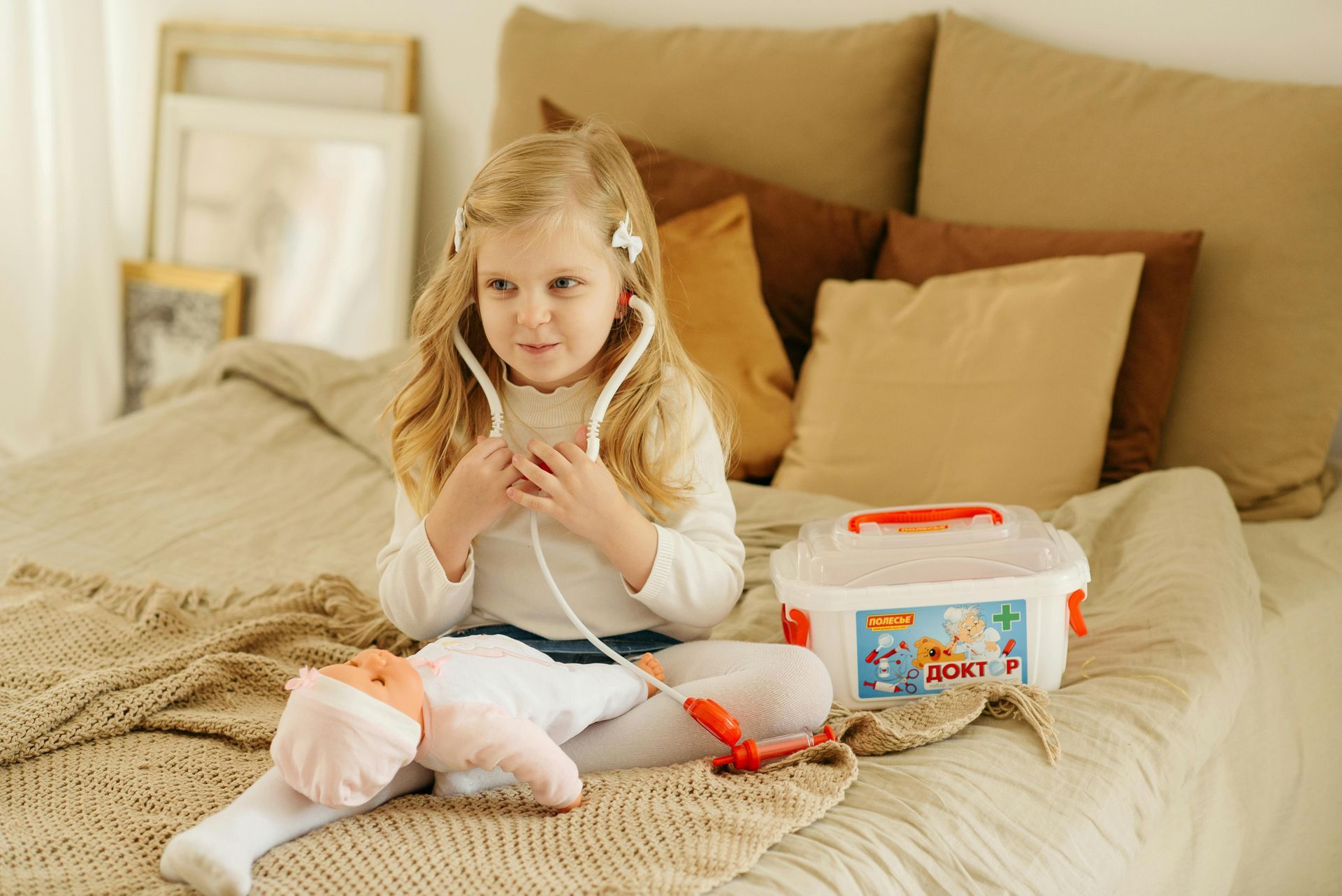 A little girl is sitting on a bed playing with a doll and a stethoscope.