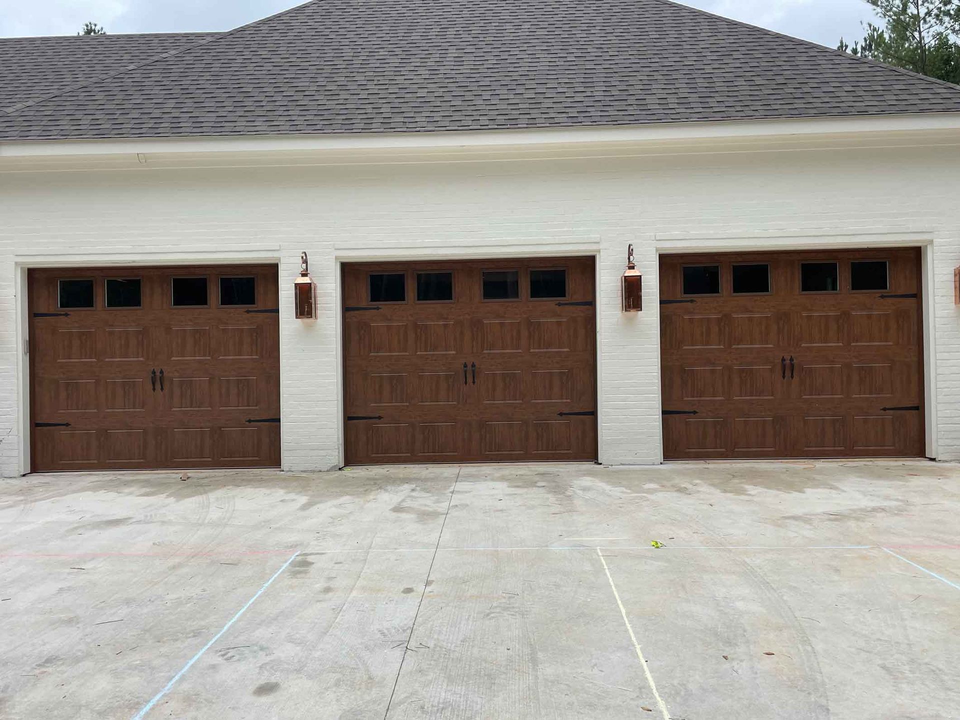Three brown garage doors on a white building with a gray roof and concrete driveway.