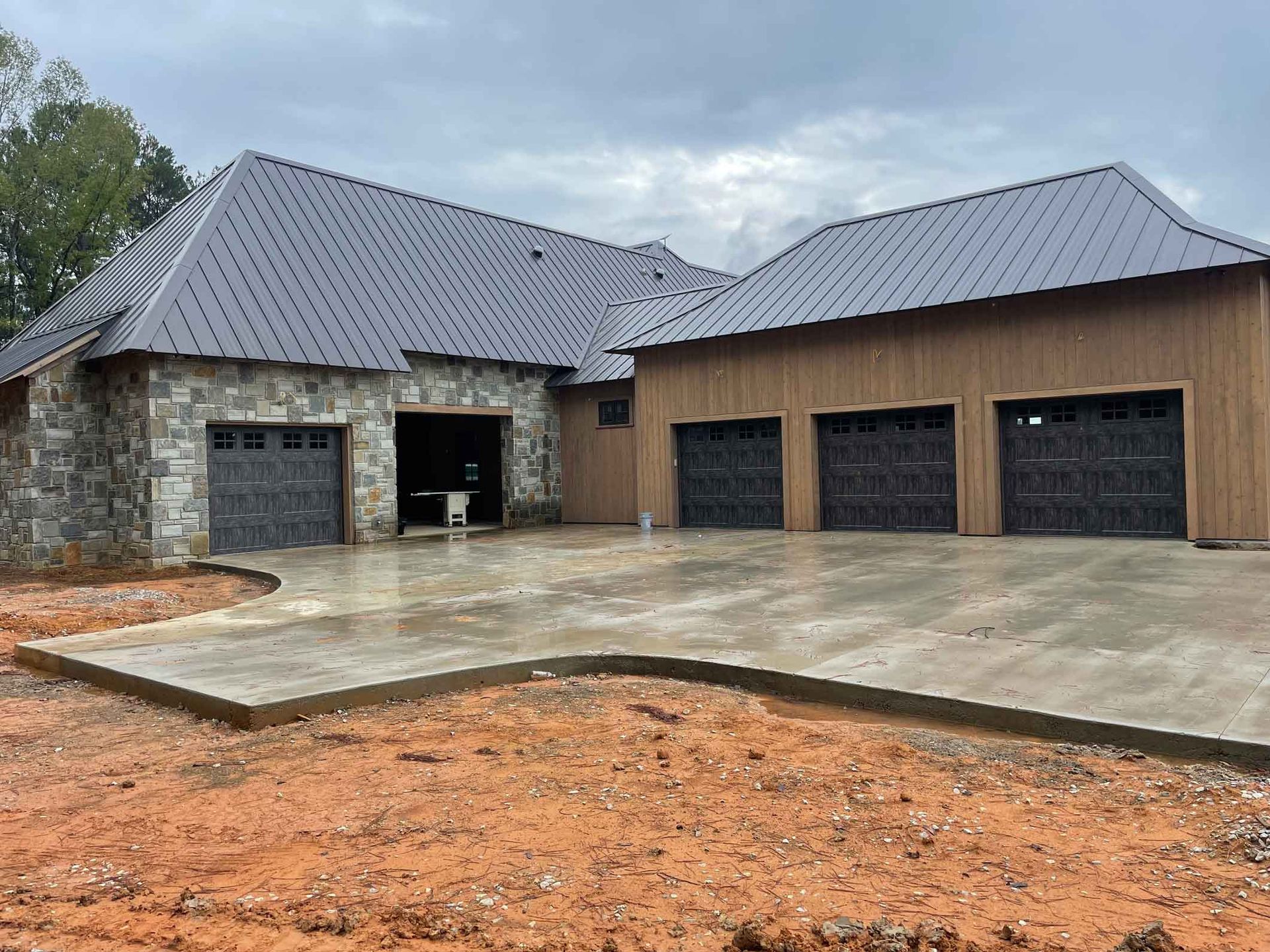 A partially built house with a concrete driveway, metal roof, and stone and brown siding, with garage doors.