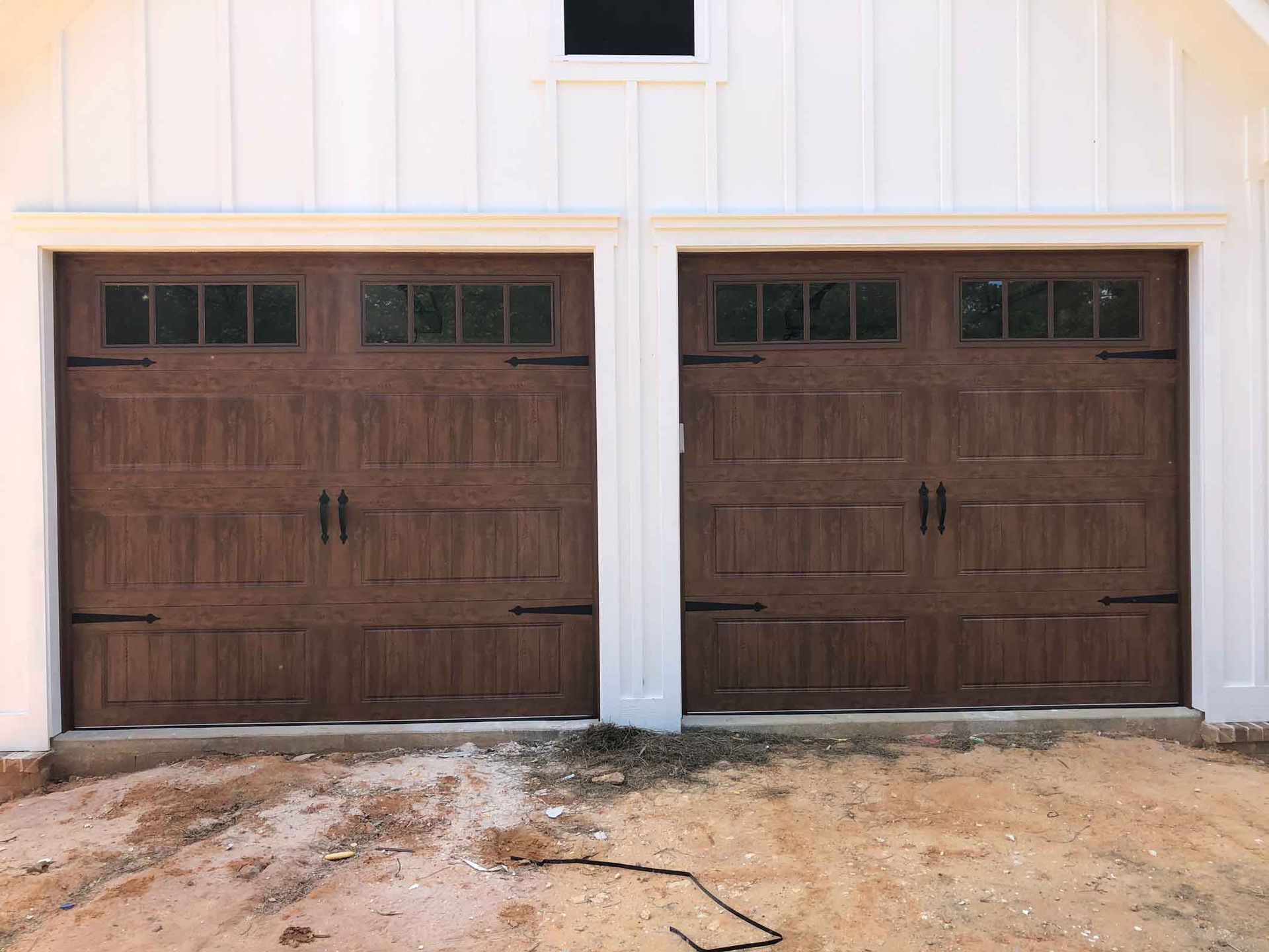 Two brown garage doors with rectangular windows, white trim, and a dirt yard.