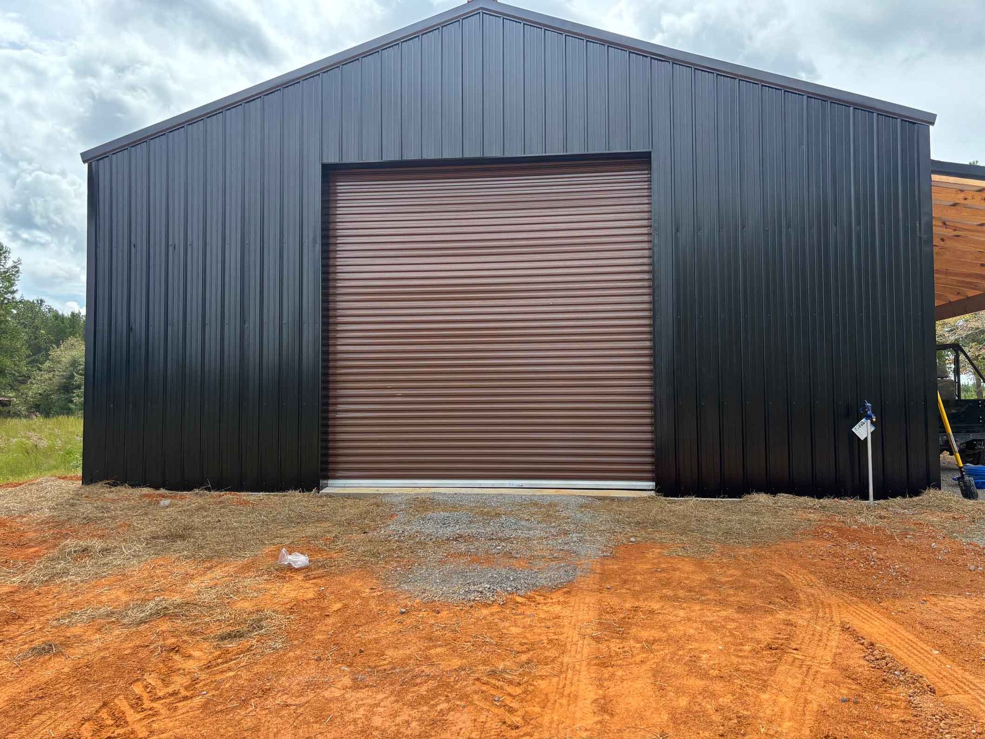 Black metal barn with a brown garage door on a dirt lot.
