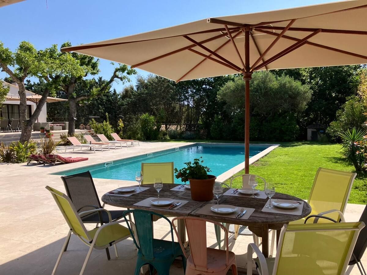 Table à manger extérieure installée sous un parasol, à côté d'une piscine et d'une pelouse verte.