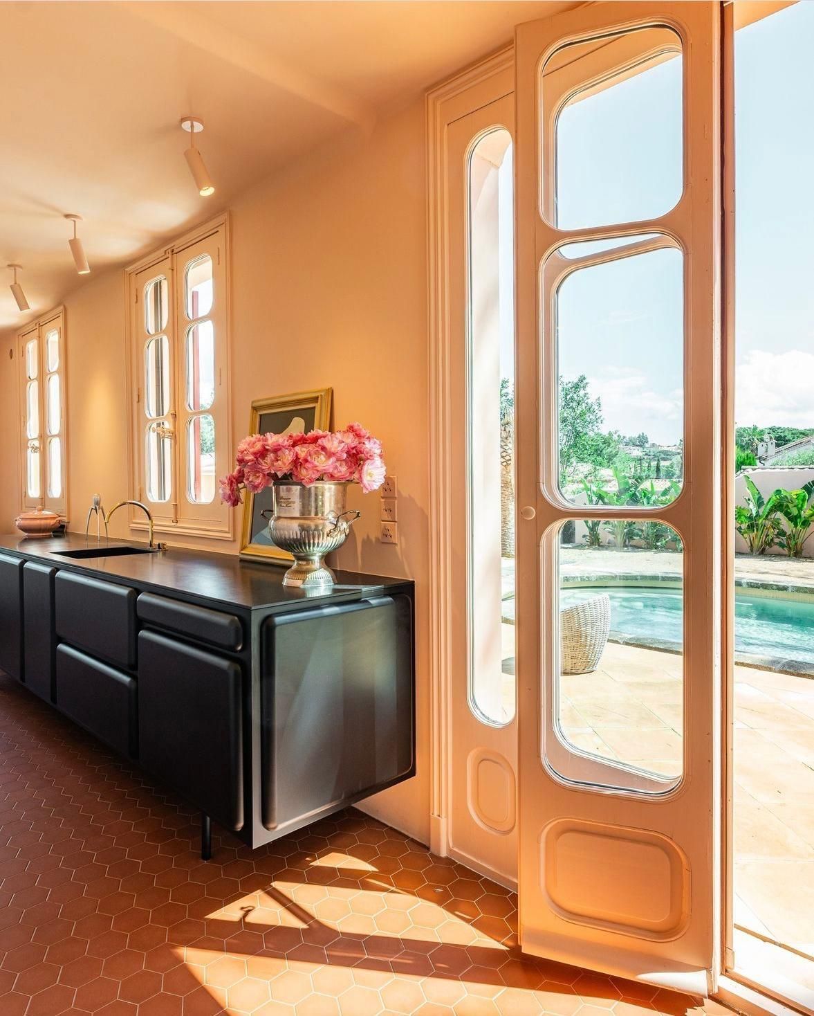 Cuisine avec placards noirs, porte ouverte sur la piscine. Fleurs roses dans un vase argenté. Lumière ensoleillée.