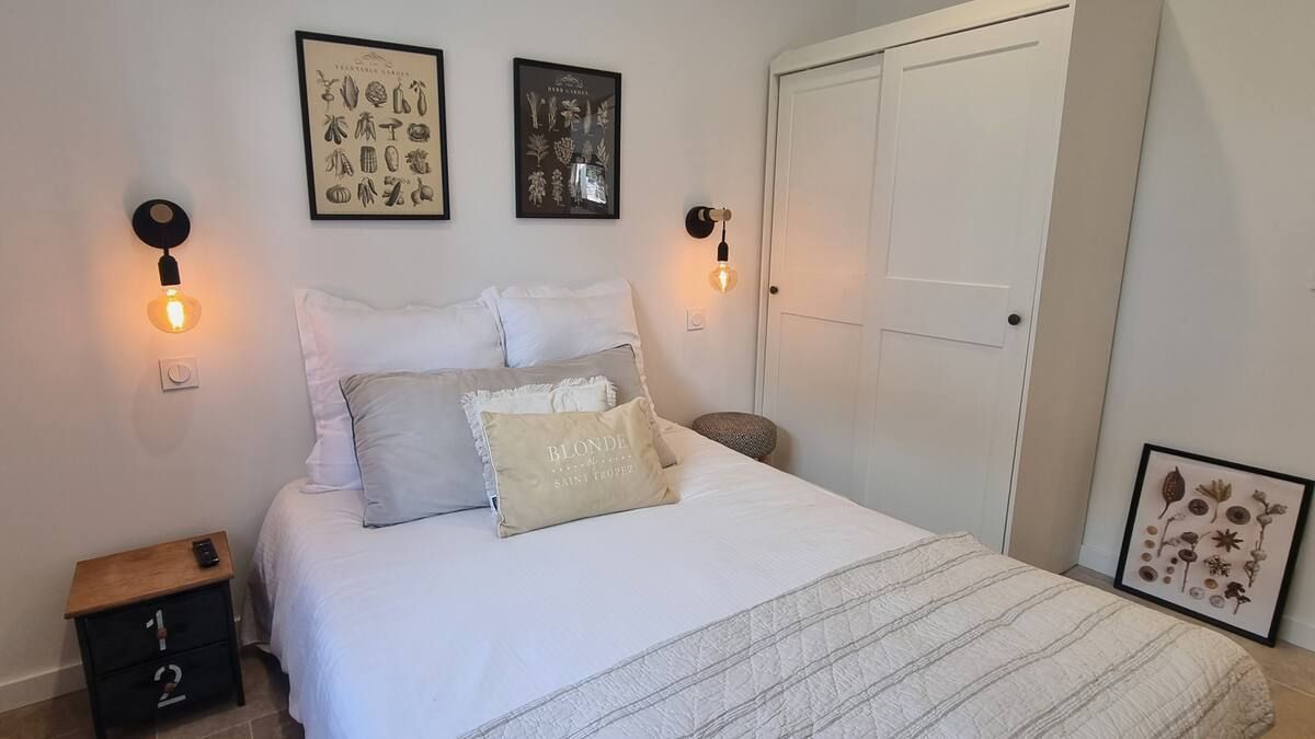 Chambre avec literie blanche, deux appliques murales, une table de chevet et des œuvres d'art encadrées.