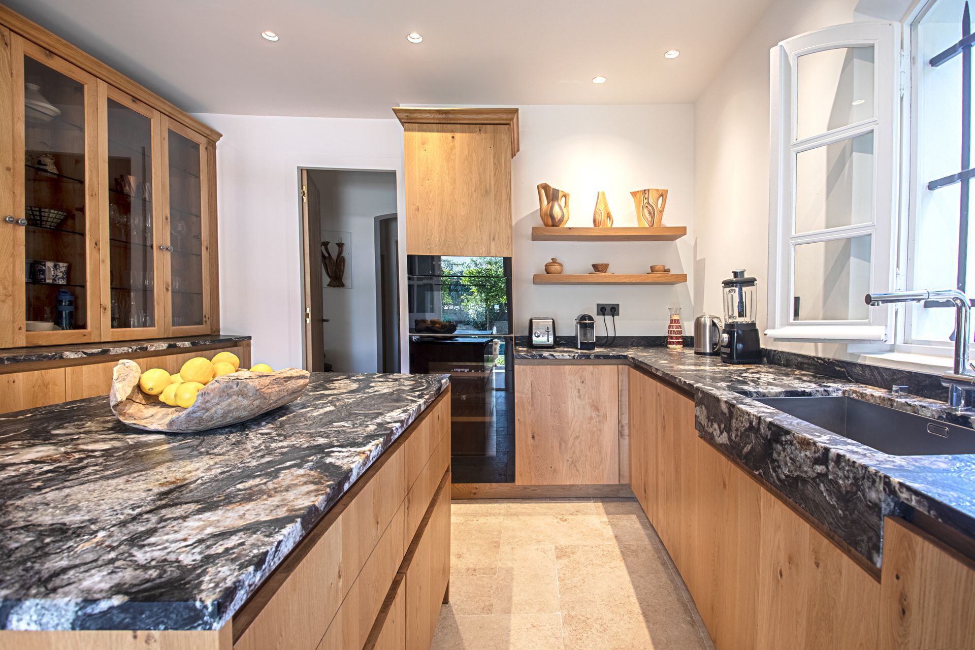 Cuisine avec armoires en bois, comptoirs en granit et vue à travers une porte et une fenêtre.