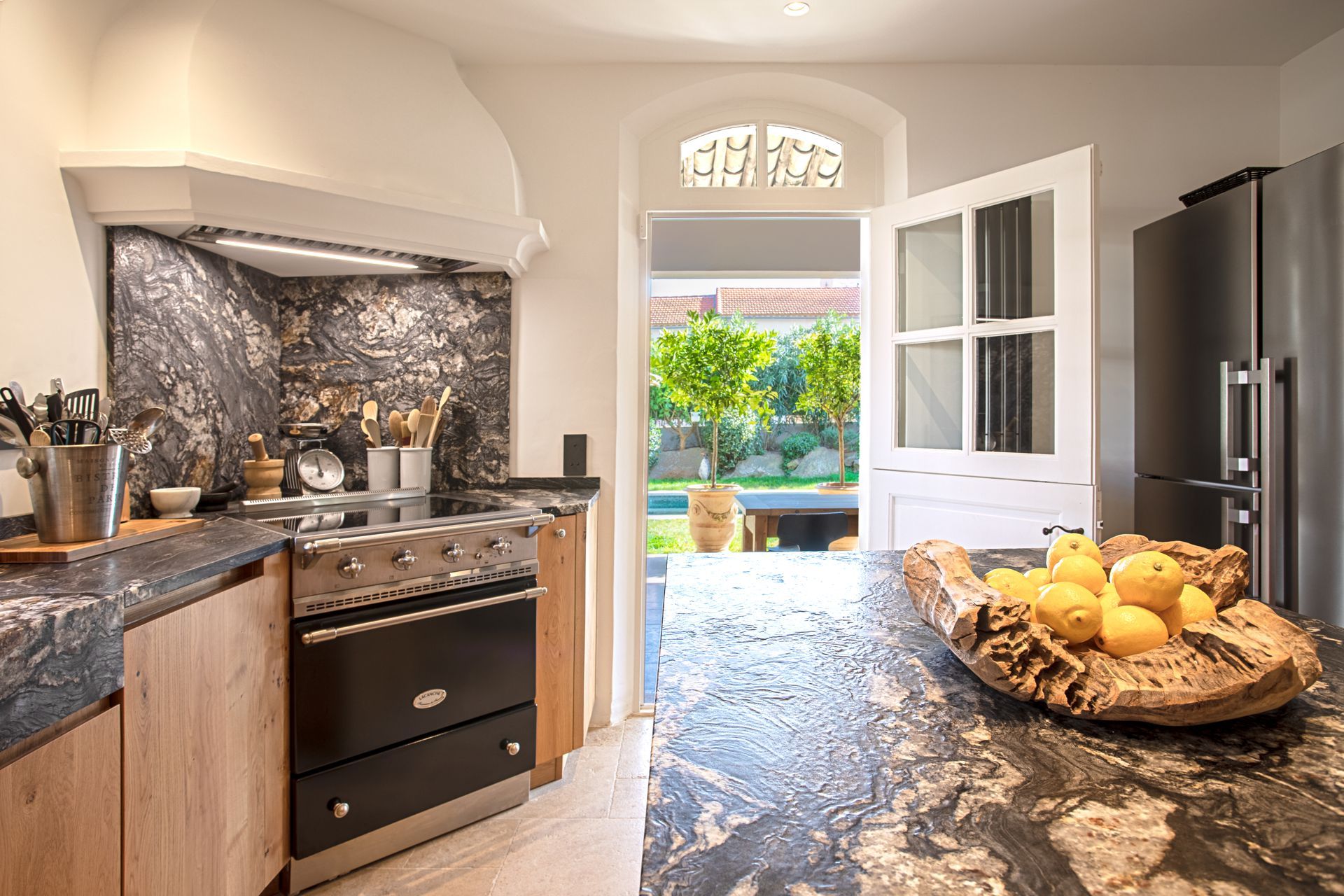 Cuisine avec accents noirs et bois, cuisinière, porte ouverte sur le jardin avec fruits jaunes.