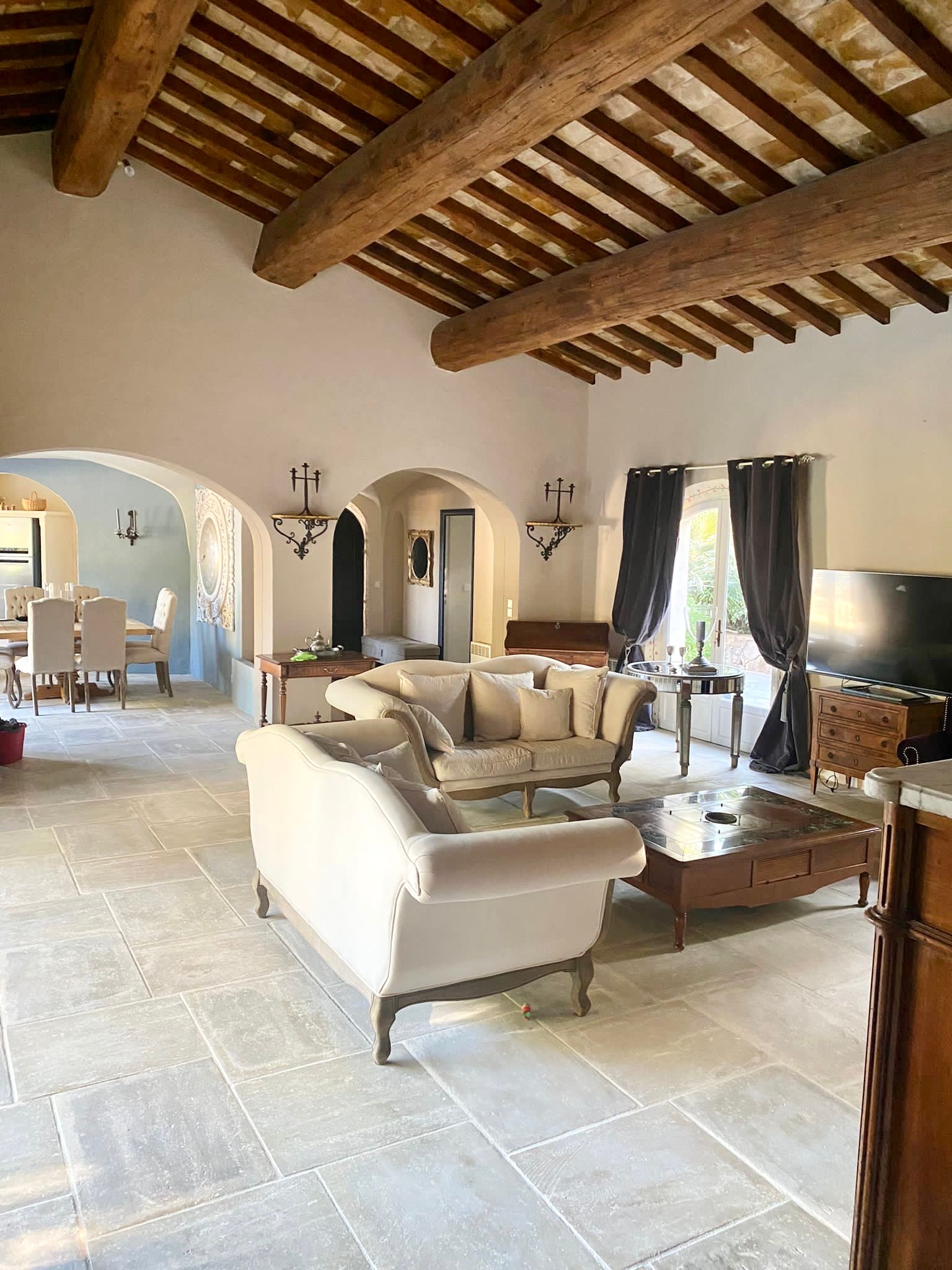 Salon spacieux avec poutres apparentes, portes cintrées et meubles aux tons neutres.