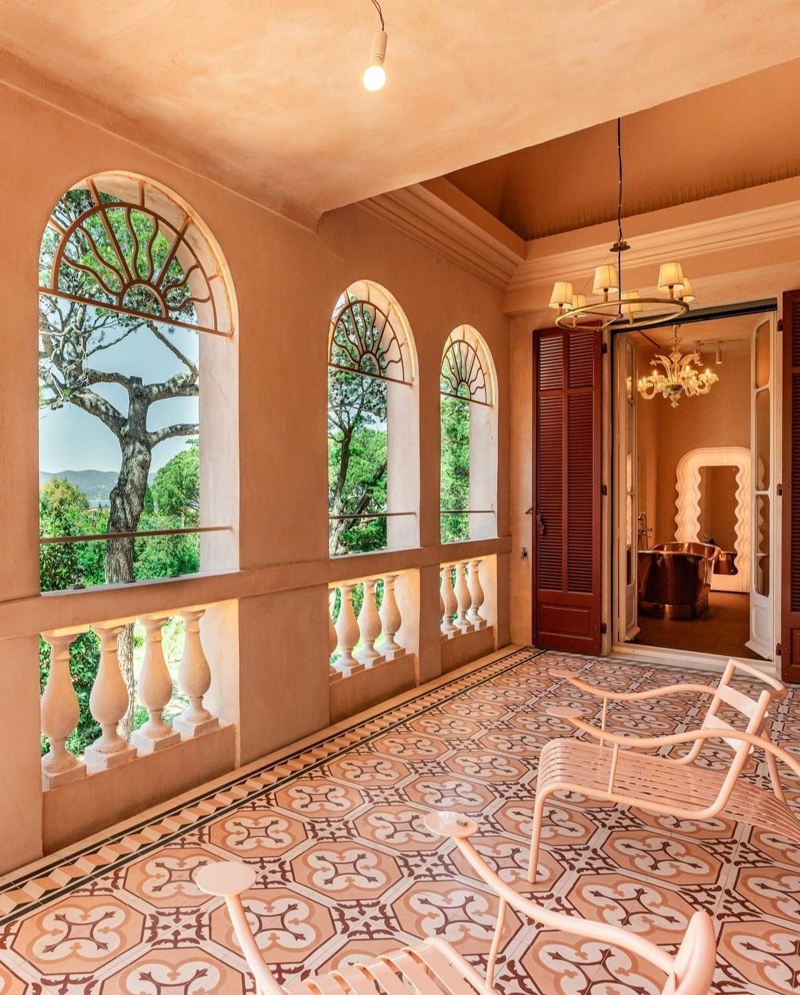 Balcon avec fenêtres cintrées donnant sur la verdure. Chaises roses sur un sol carrelé à motifs. Porte ouverte sur l'intérieur.