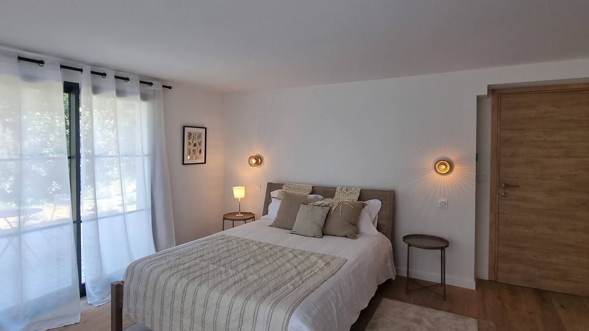 Chambre avec lit, deux tables de chevet, deux appliques murales, fenêtre avec rideaux transparents et une porte en bois.