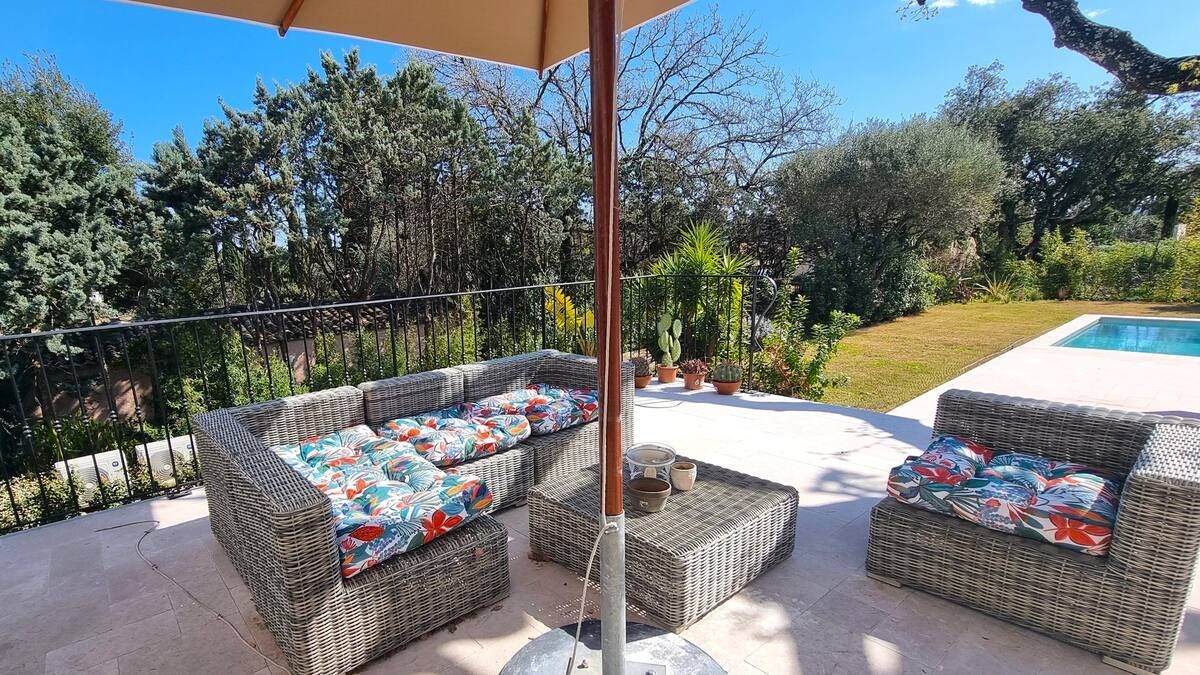 Terrasse extérieure avec mobilier en osier et coussins fleuris, parasol et piscine en arrière-plan.