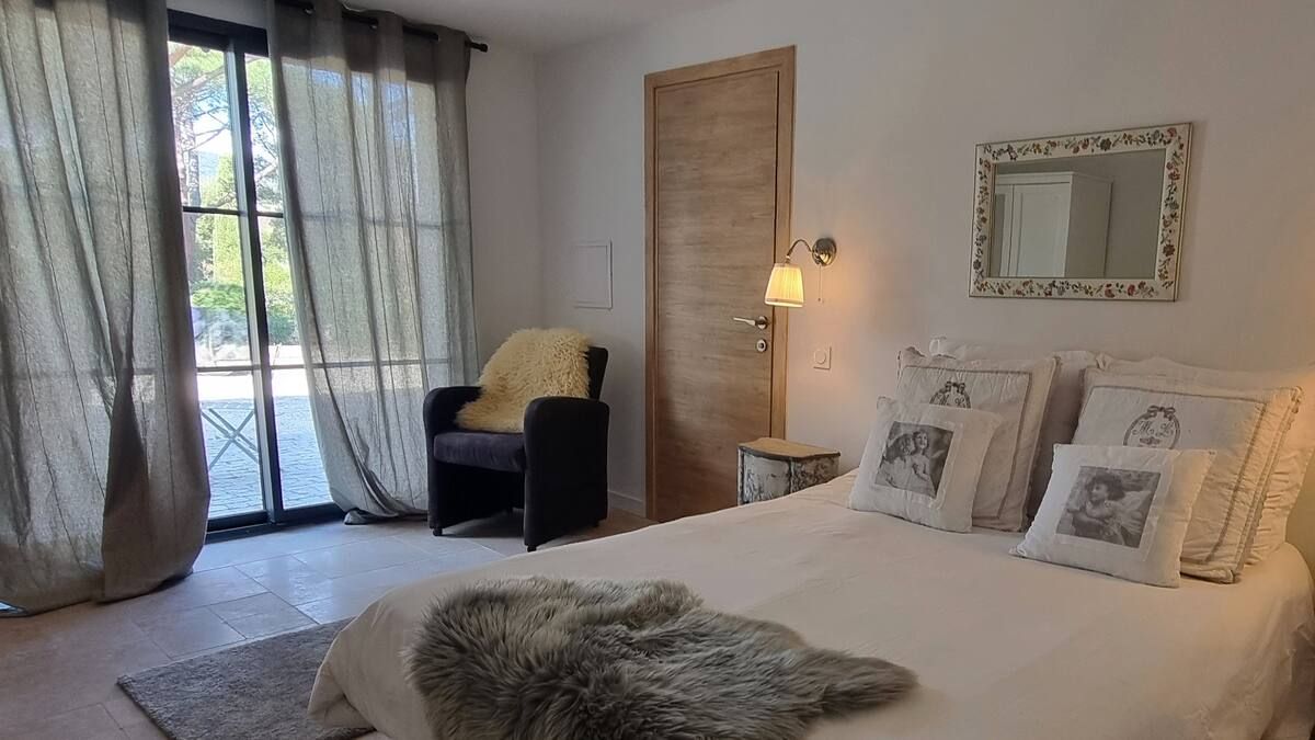 Chambre avec lit, fauteuil, fenêtre, rideaux et miroir. Murs blancs et palette de couleurs neutres.