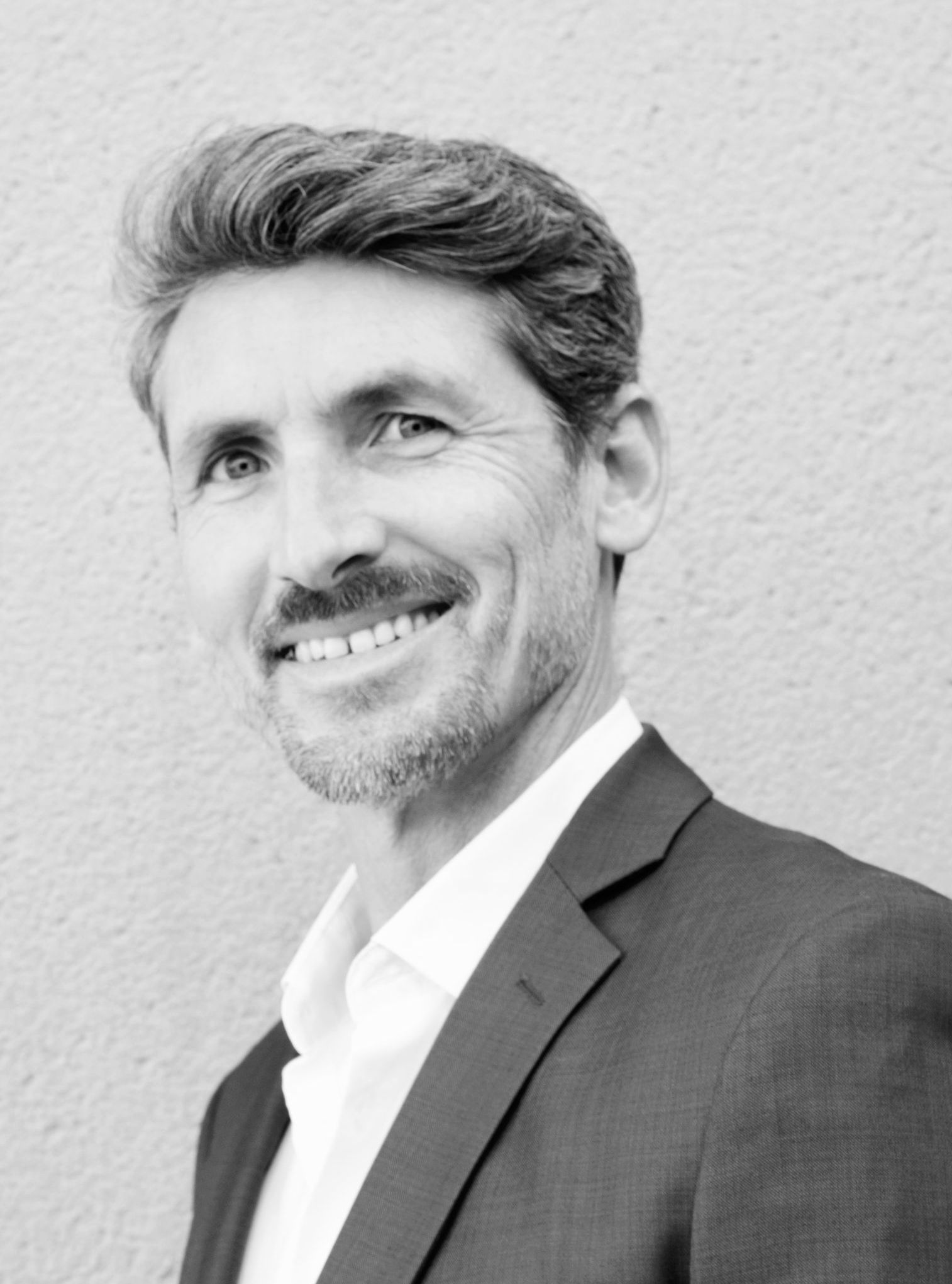 Homme aux cheveux poivre et sel, vêtu d'un costume, souriant, debout à l'extérieur près d'un mur beige.