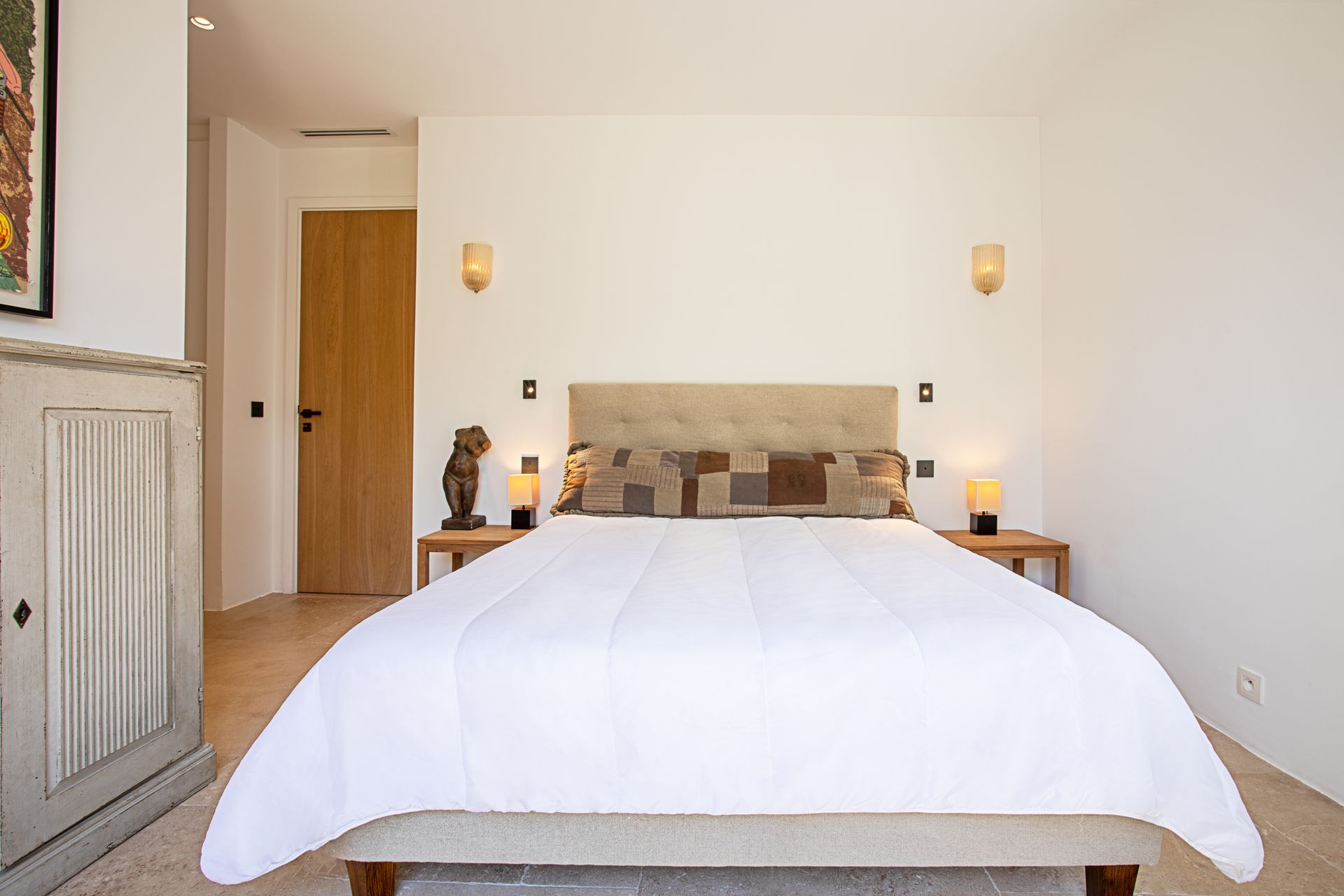 Chambre avec lit, tête de lit, tables de chevet, lampes et œuvres d'art. Murs blancs, sol clair, porte en bois.