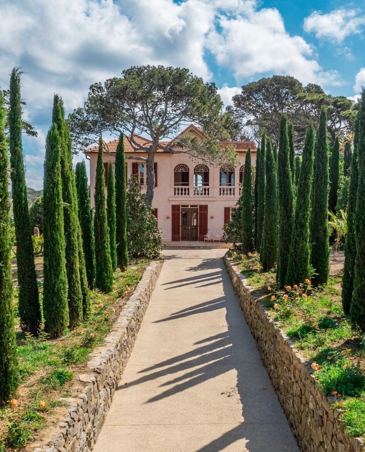Chemin menant à une maison rose bordée de cyprès, de murs en pierre et sous un ciel bleu.