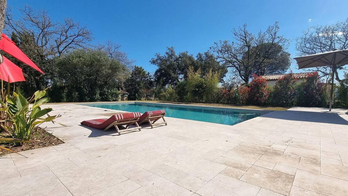 Piscine avec chaises longues sous un ciel bleu. Parasols rouges, arbres et verdure tout autour.