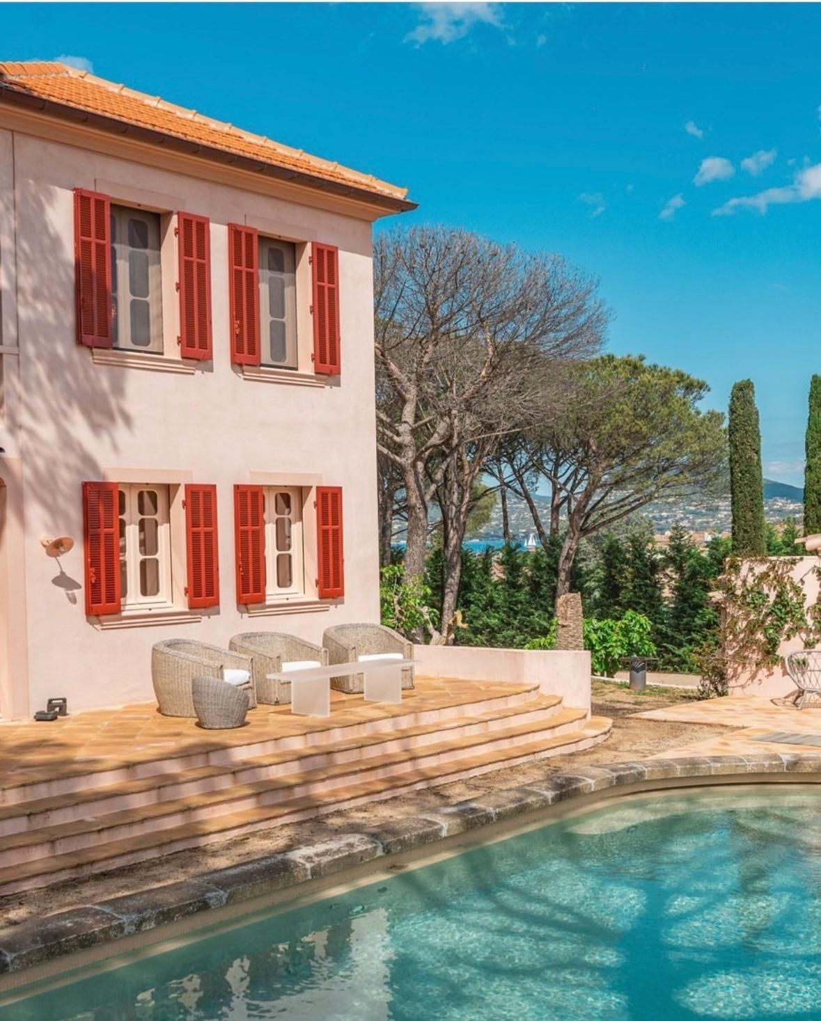 Maison en stuc rose avec volets rouges et piscine, dans un cadre ensoleillé.