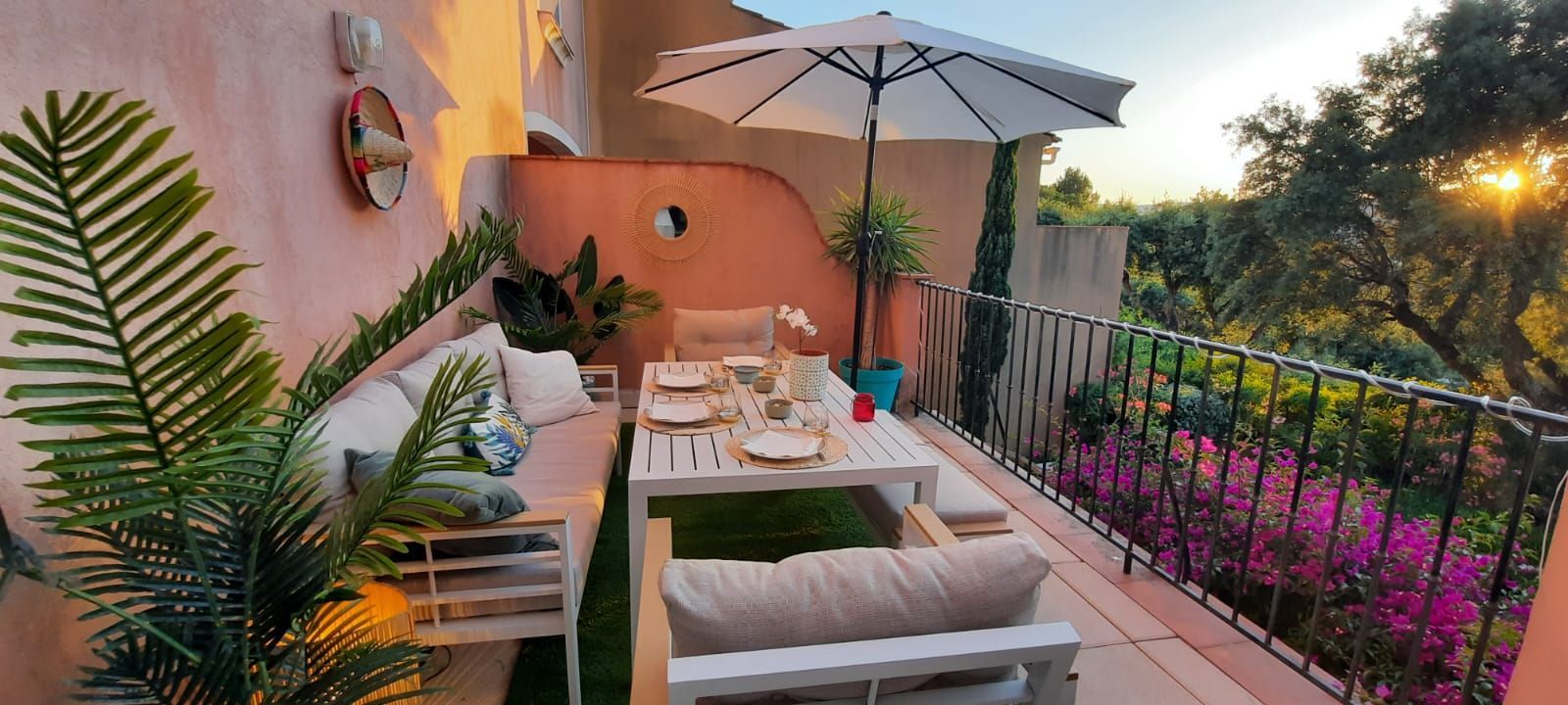Terrasse extérieure avec mobilier blanc, parasol et table dressée pour les repas, offrant une vue sur des fleurs et des arbres violets.