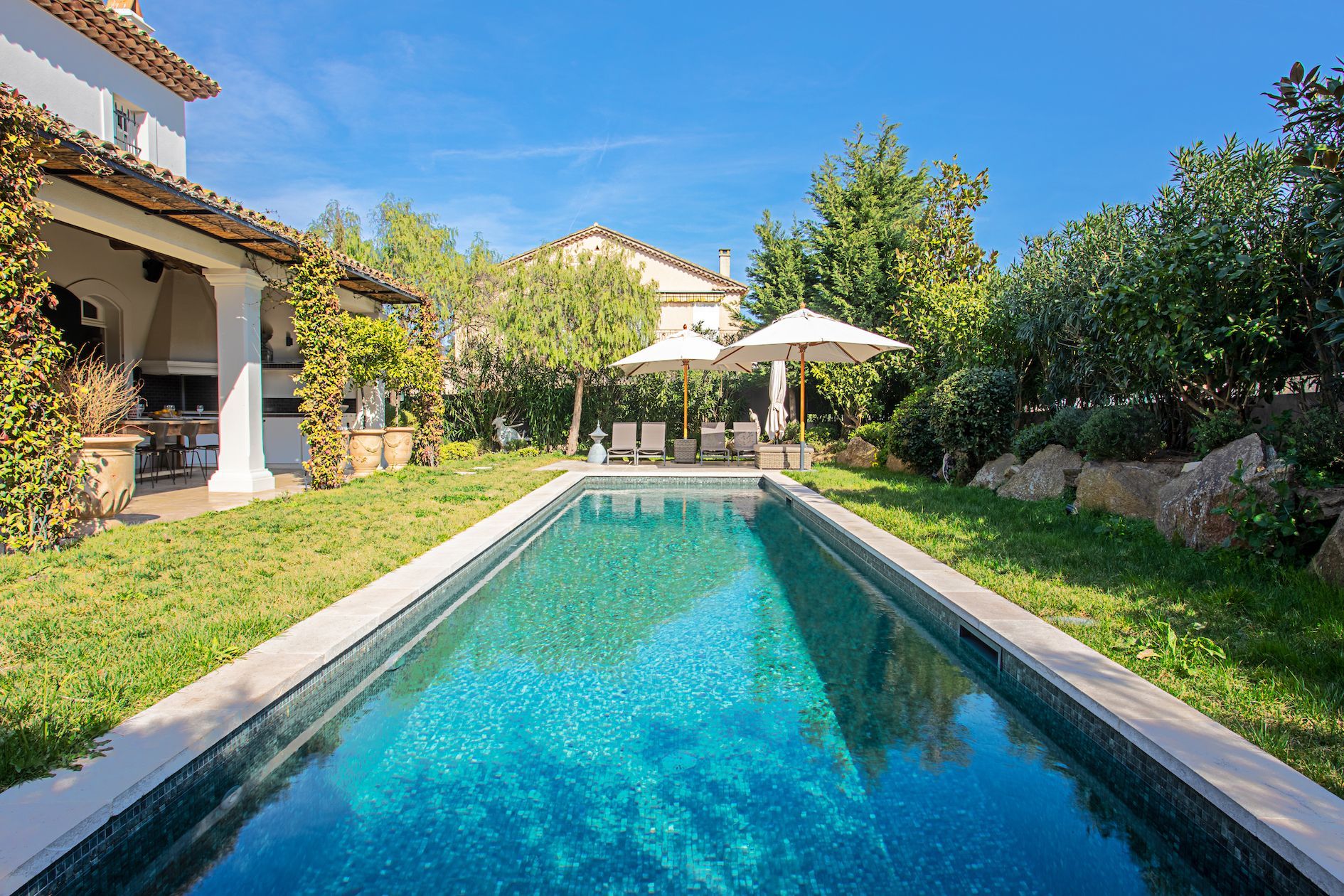 Piscine à l'eau turquoise dans une cour verdoyante, à côté d'une villa blanche sous un ciel bleu.