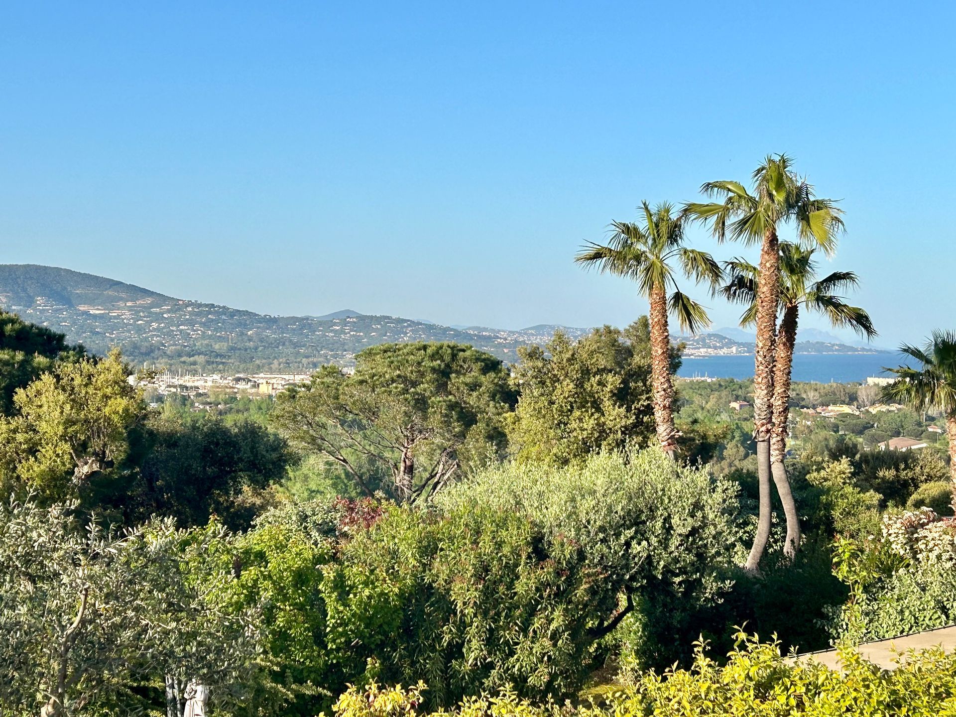 Coin repas extérieur avec table blanche, coussins et verdure. Murs roses, parasol, ensoleillé.