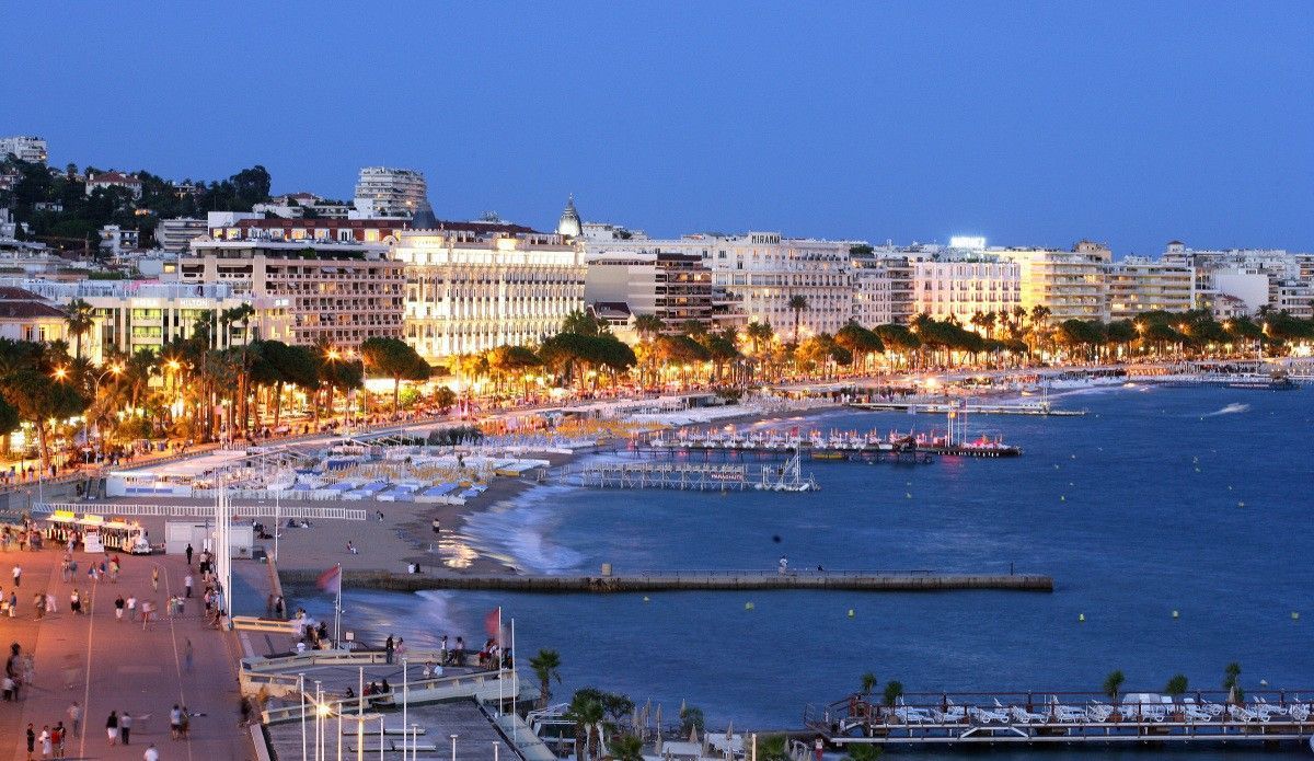 Ville côtière au crépuscule, avec ses bâtiments illuminés, sa plage, son port et ses eaux calmes.
