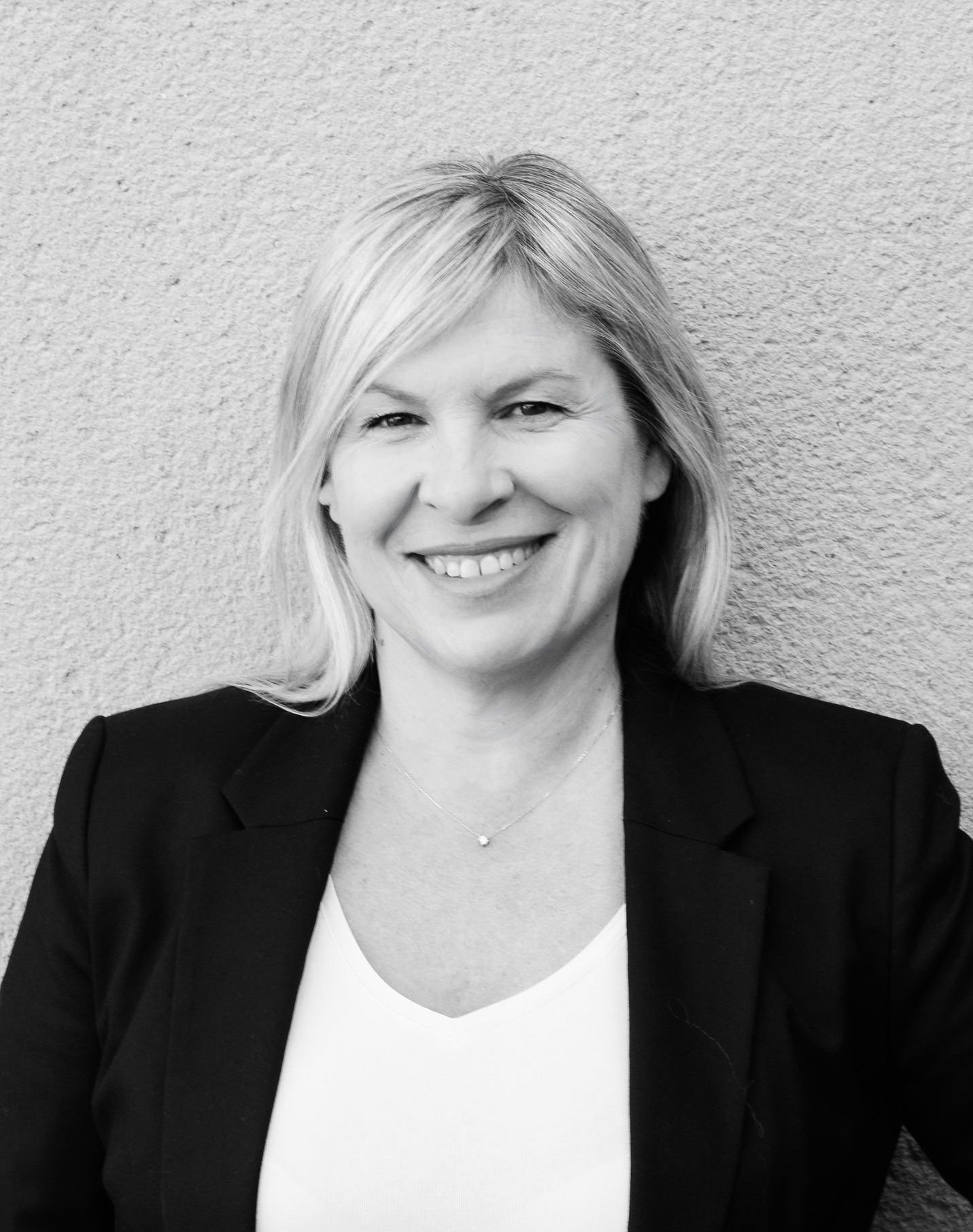 Une femme blonde en blazer noir sourit à l'objectif, posant devant un mur neutre.