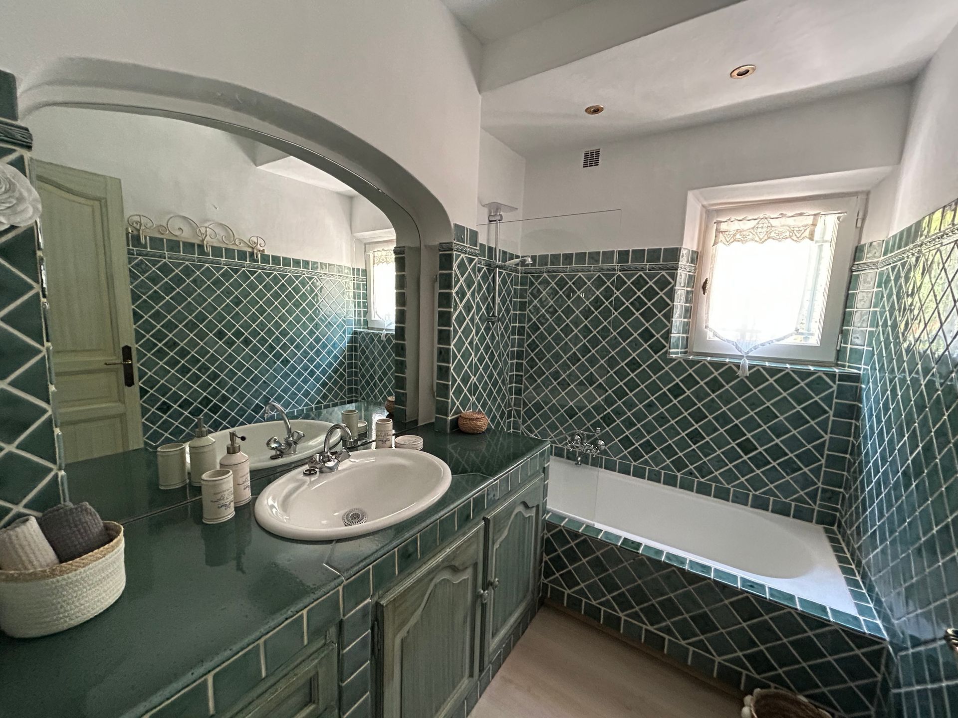 Salle de bain carrelée de vert avec meuble-lavabo, miroir et baignoire. Lumière du jour par une petite fenêtre.