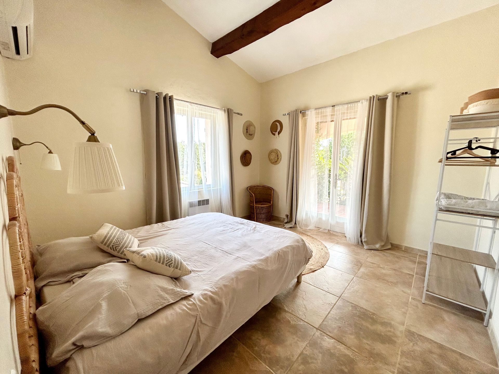 Chambre avec lit, fenêtres avec rideaux et étagère. Tons beiges, lumière naturelle.