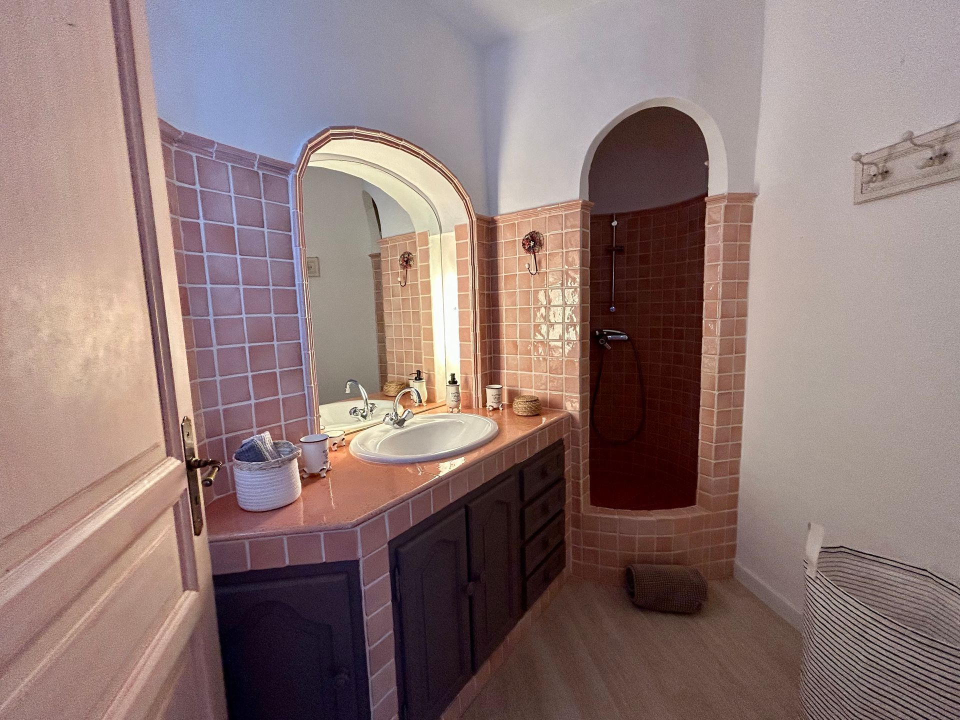 Salle de bain avec murs carrelés roses, armoires sombres et miroir arqué.