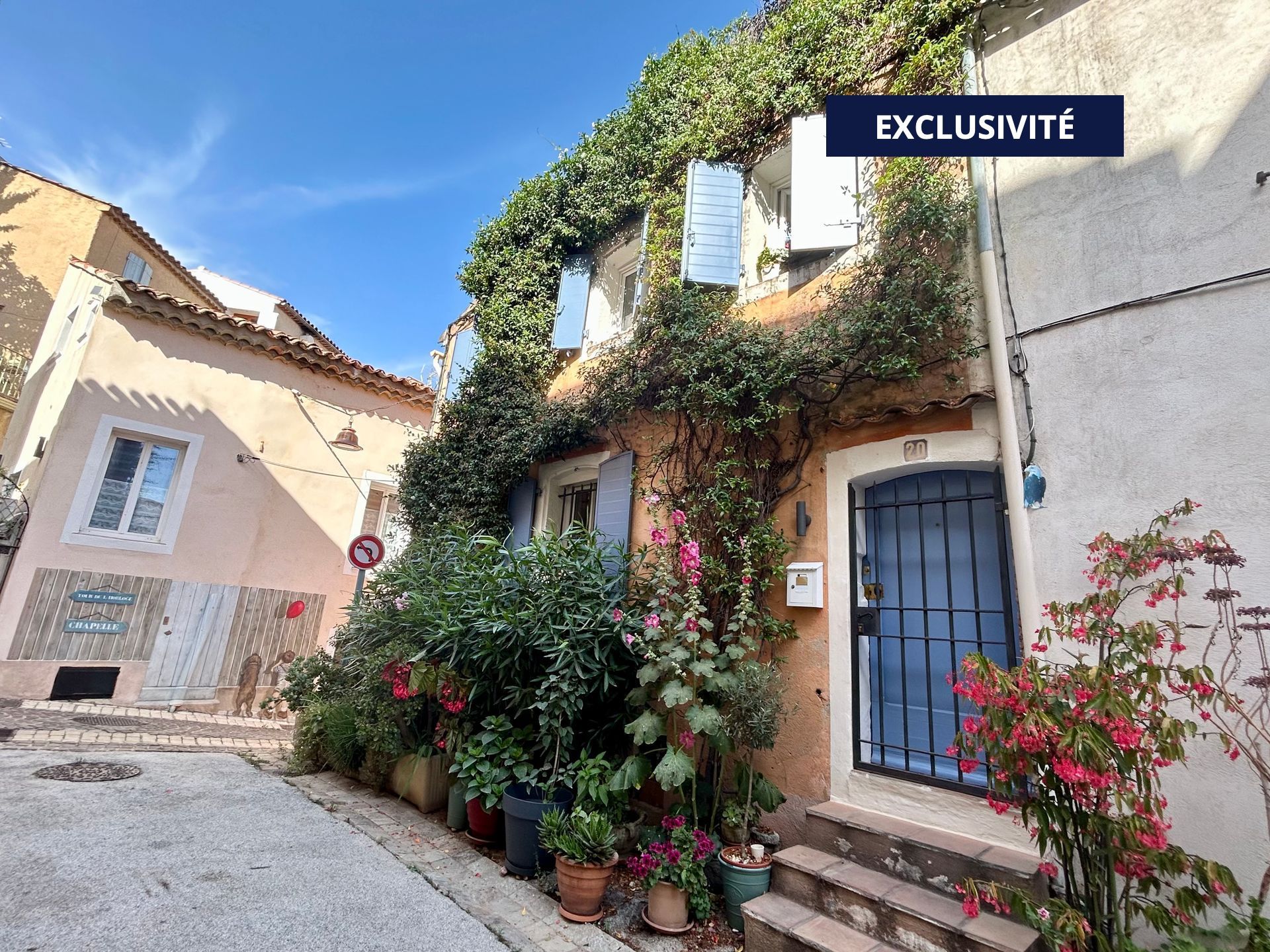 Rue pavée étroite bordée de bâtiments colorés et de verdure, inscription « VENDU PAR NOUS », sceau « EXCLUSIF ».