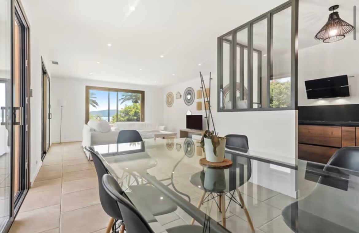 Salle à manger avec table en verre, chaises noires et une fenêtre donnant sur l'océan.