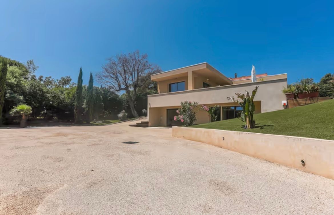 Maison moderne à deux étages avec murs beiges, allée en gravier et pelouse verte sous un ciel bleu clair.