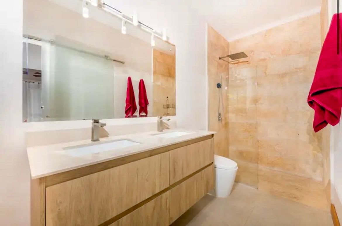 Salle de bain moderne avec vanité en bois, grand miroir, douche et serviettes rouges.