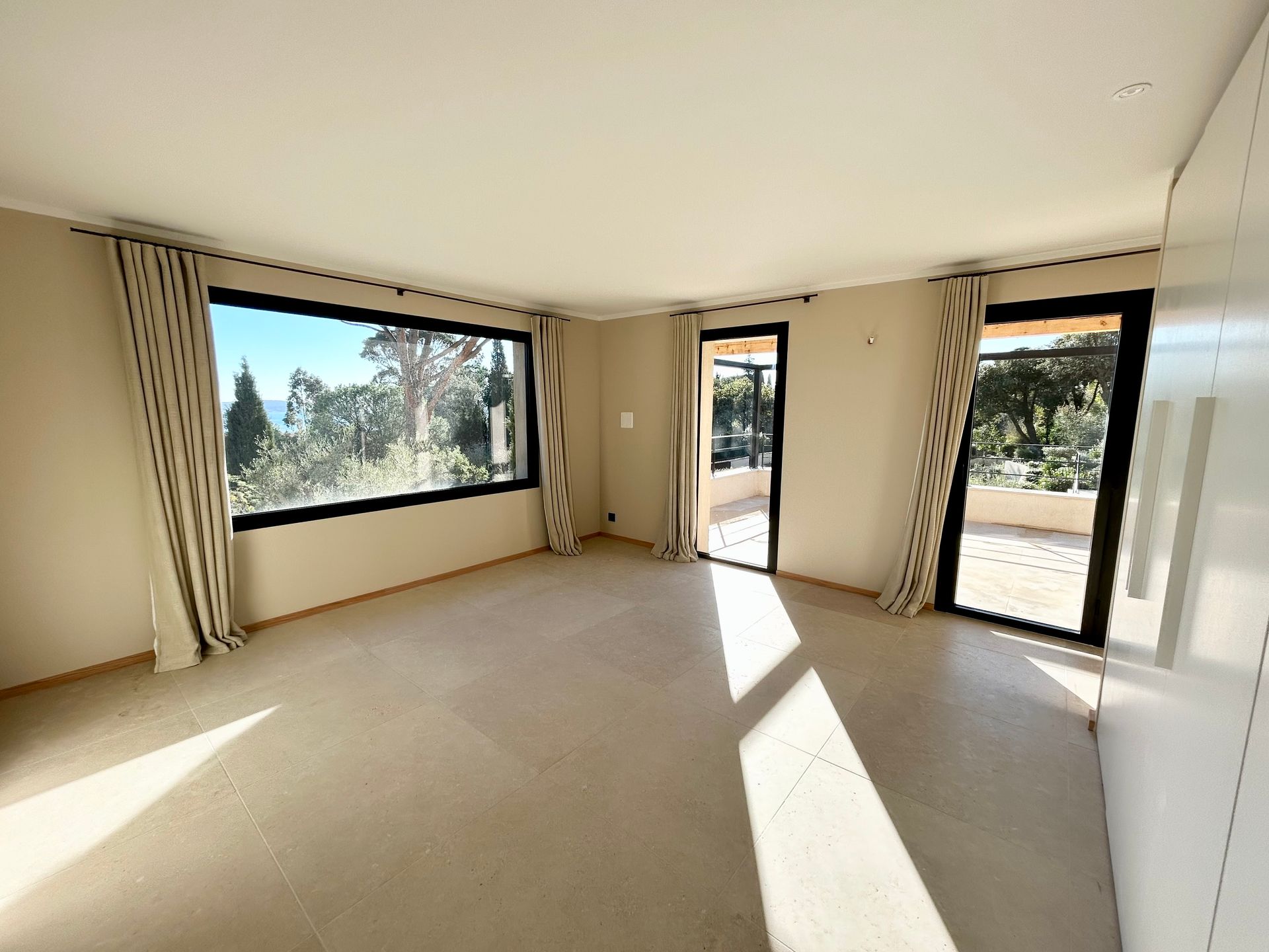 Chambre vide avec de grandes fenêtres offrant une vue panoramique et porte coulissante donnant sur un patio, tons neutres.