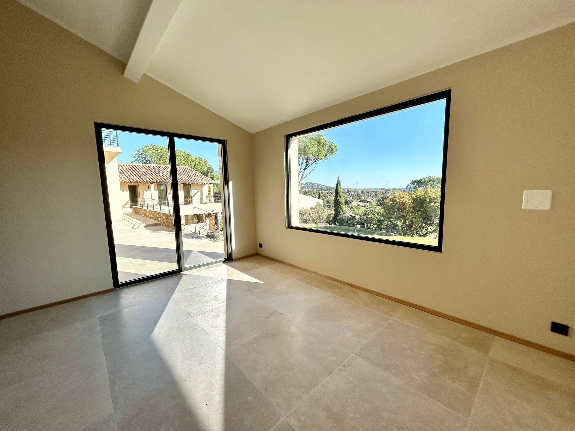 Chambre vide avec de grandes fenêtres, de la lumière du soleil et une vue panoramique sur un bâtiment et un paysage.