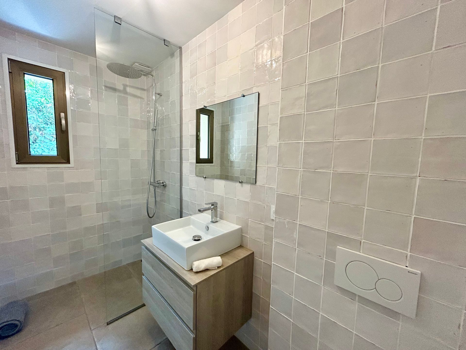 Salle de bain avec murs carrelés gris, une vanité en bois et une fenêtre avec vue extérieure.