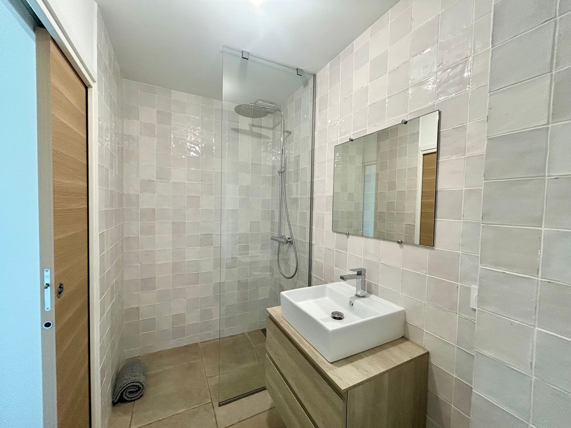 Salle de bain avec lavabo blanc, douche en verre et carrelage carré clair. Meuble-lavabo et porte en bois.