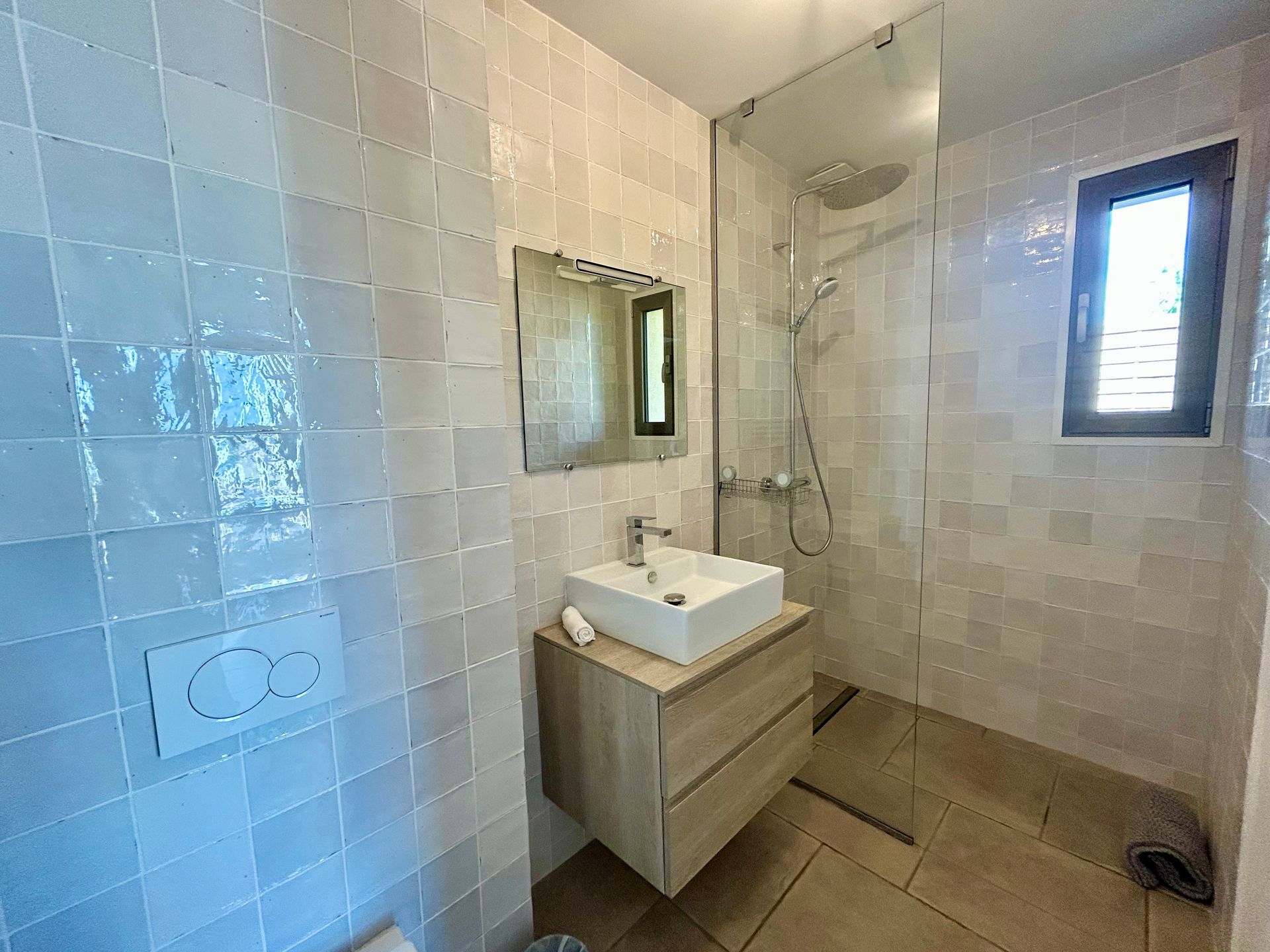 Salle de bain avec murs en carrelage blanc, vanité en bois, douche et une petite fenêtre.