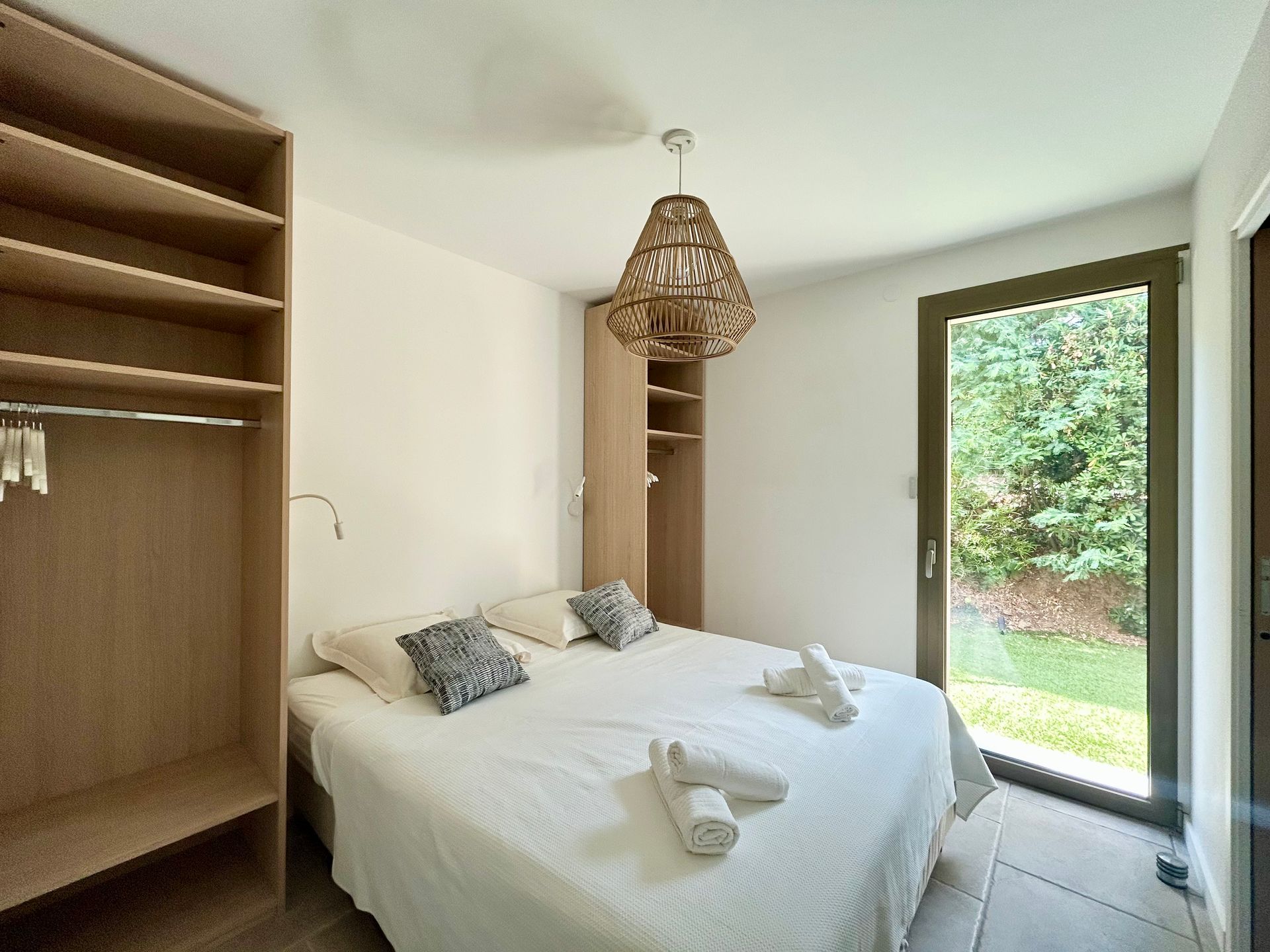 Chambre avec lit, placard et grande fenêtre donnant sur un feuillage vert. Une lampe en bois est suspendue au plafond.