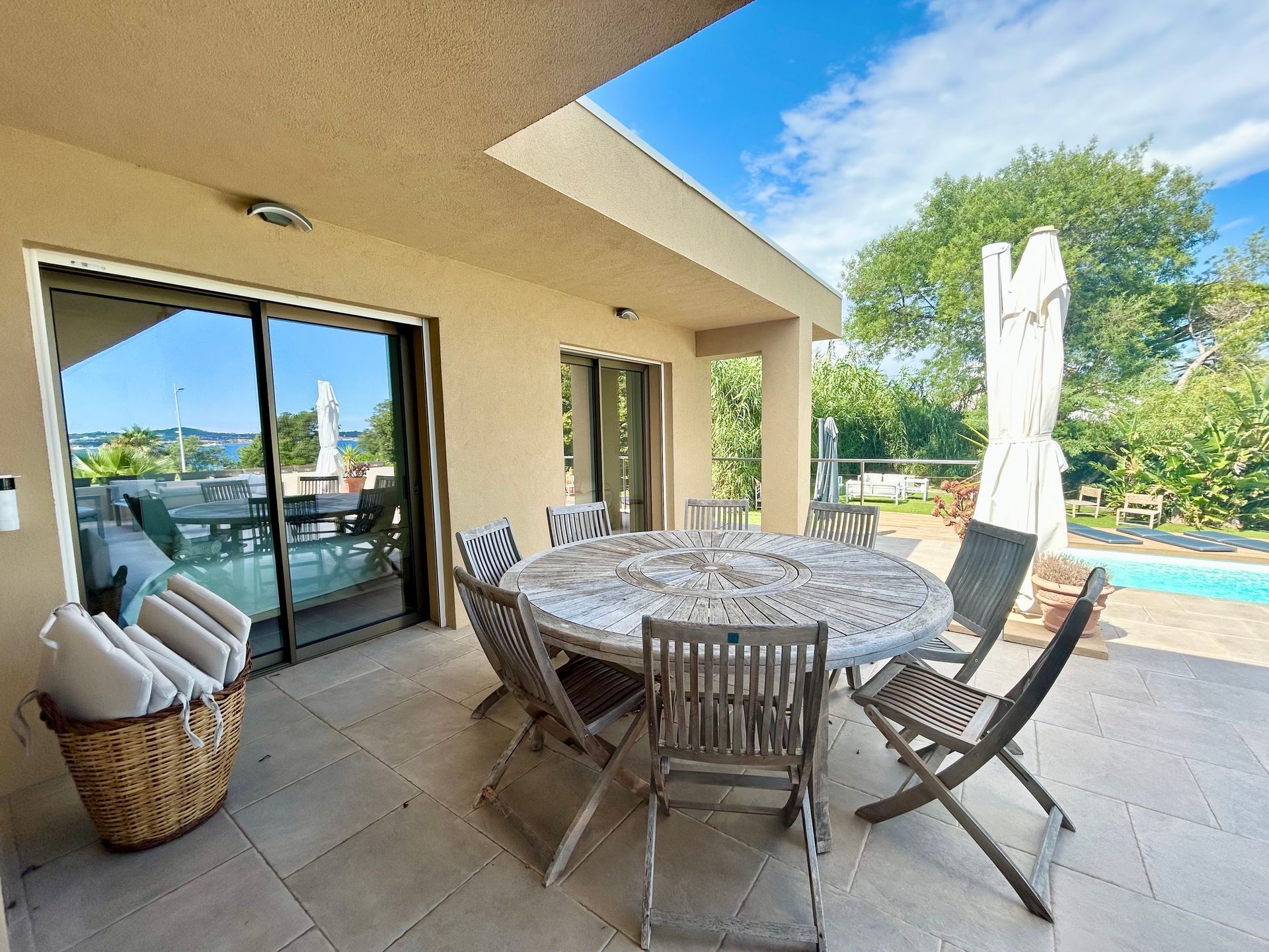Patio avec table ronde et chaises, donnant sur la piscine et la cour; bâtiment de couleur claire et ciel ensoleillé.