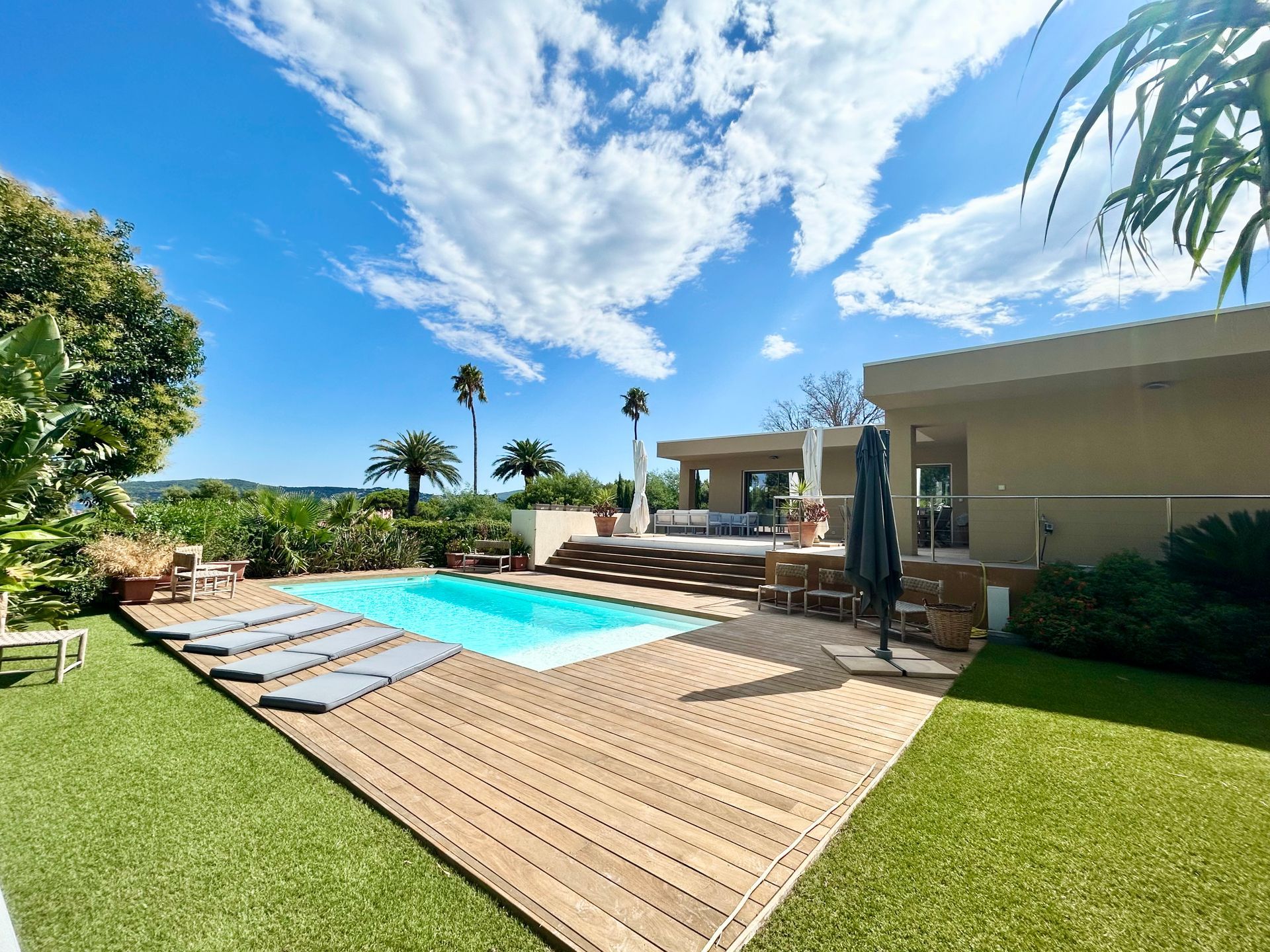 Une scène extérieure ensoleillée avec une piscine, un bâtiment sous un ciel bleu avec des nuages.