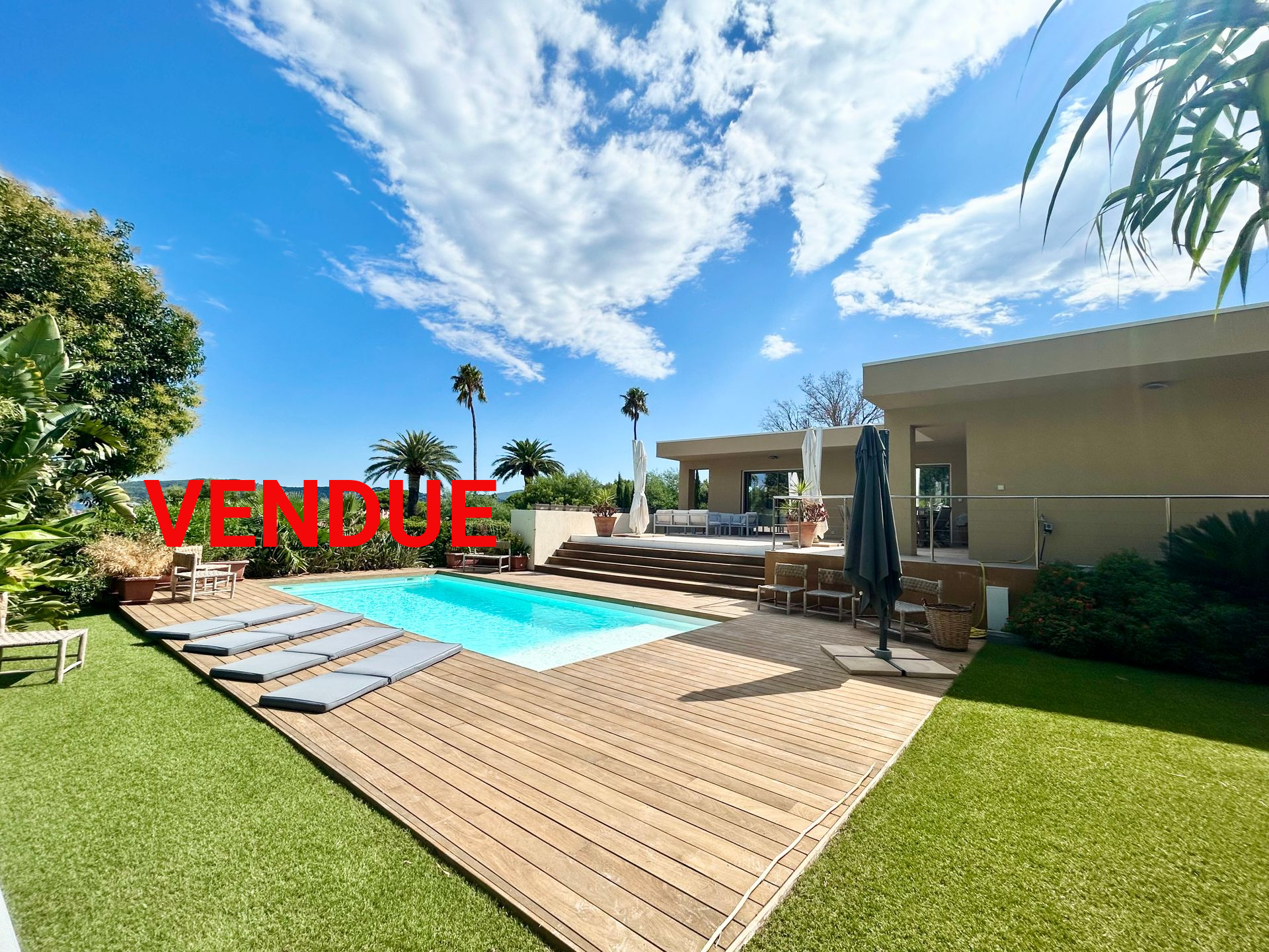Jardin avec piscine, terrasse et pelouse, sous un ciel bleu avec un panneau « À VENDRE ».