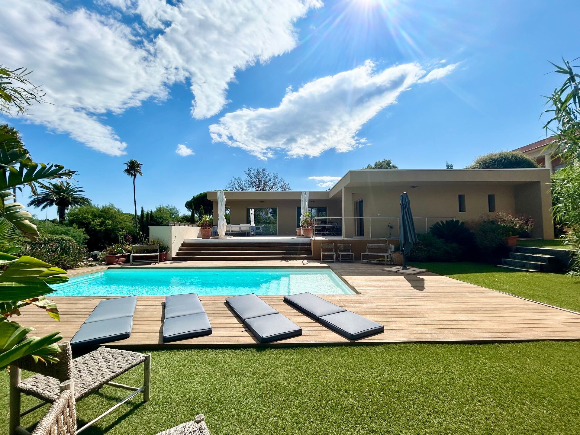 Maison avec piscine et chaises longues par une journée ensoleillée. Ciel bleu avec nuages, pelouse verte.