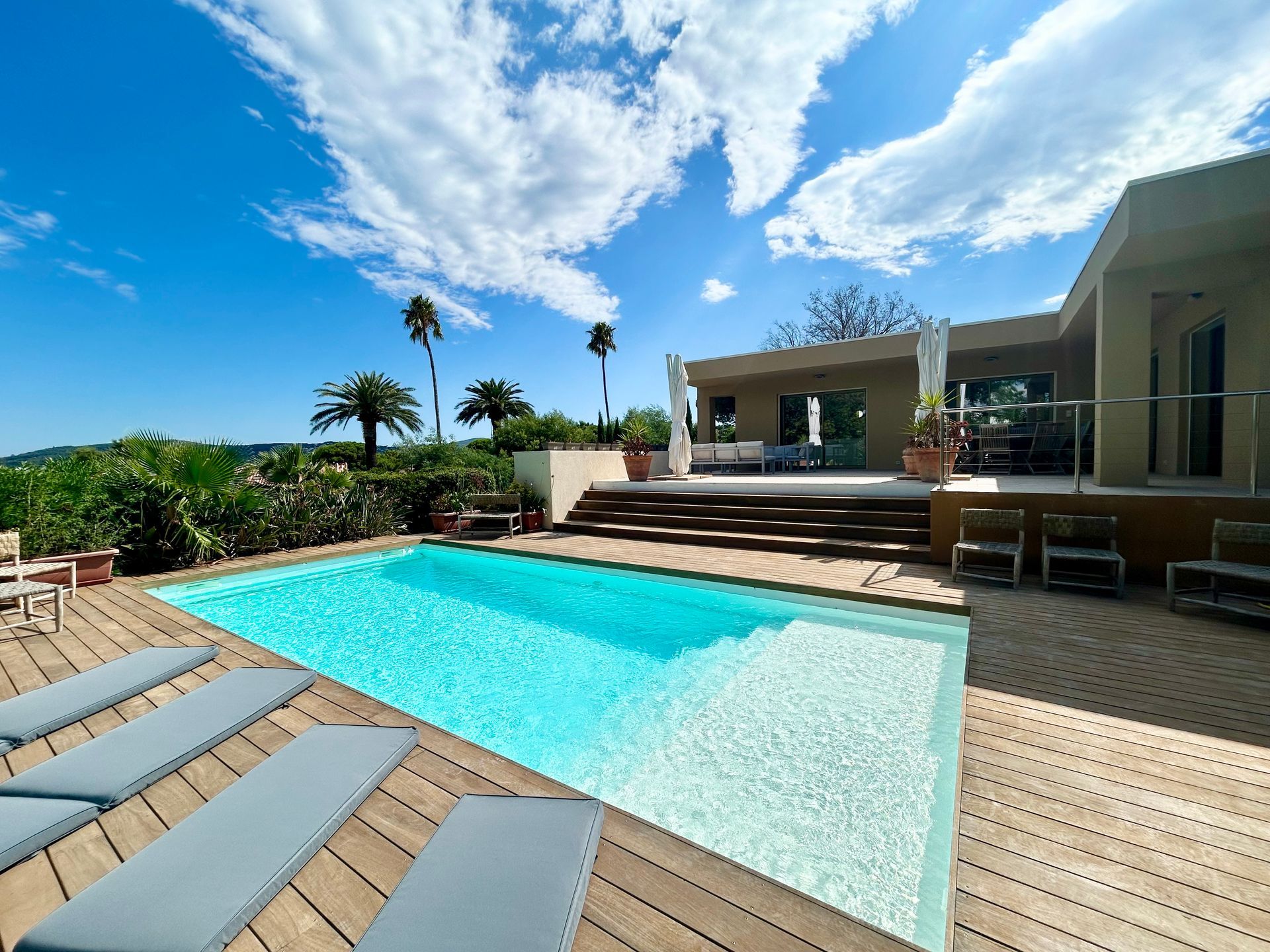Piscine avec terrasse en bois et transats, villa en arrière plan, sous ciel bleu.
