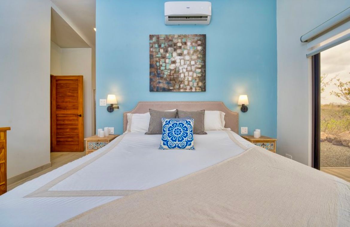 Chambre avec murs bleus, lit king size et œuvre d'art au-dessus de la tête de lit, porte et fenêtre en bois.