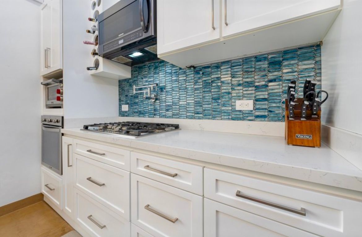 Cuisine blanche avec dosseret en mosaïque bleue, appareils électroménagers en acier inoxydable et comptoirs blancs.