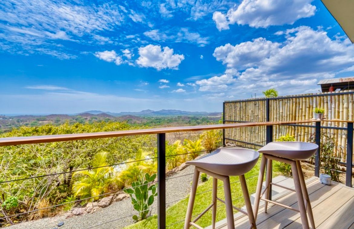 Balcon avec deux tabourets donnant sur un paysage verdoyant et des montagnes sous un ciel bleu vif.